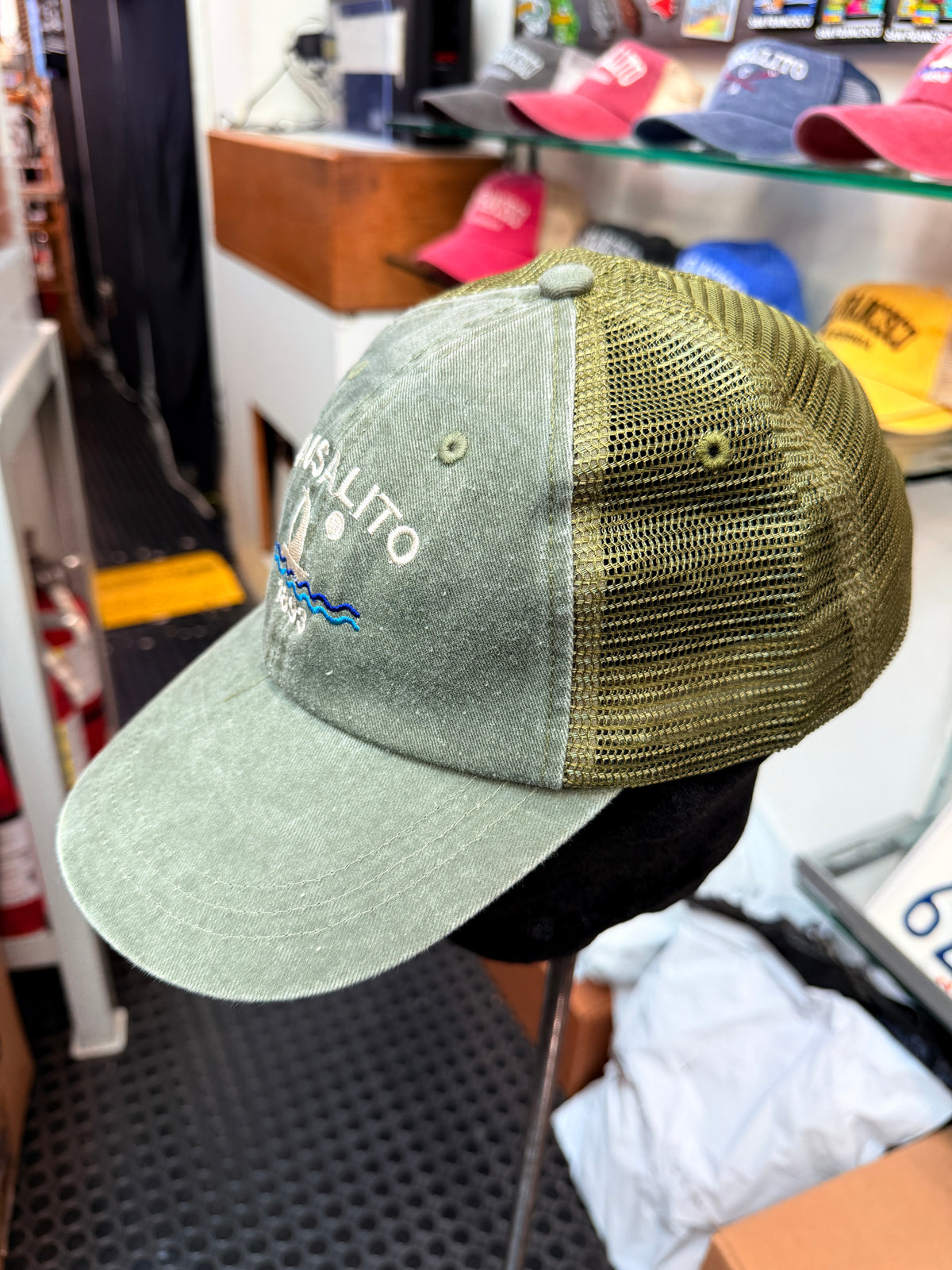 A side view of a washed, pigment-dyed sage green 100% cotton trucker baseball cap displayed on a stand in a store. The cap features a matching sage green mesh back panel, and the front panel displays white embroidered text reading "SAUSALITO" arched above a white sailboat with blue stitched waves and a small white moon.