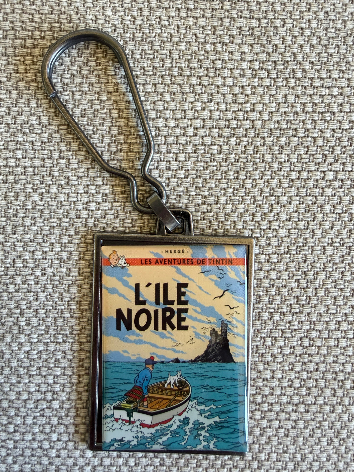 Front view of the rectangular Tintin L'Île Noire metal keychain resting on a textured, beige woven fabric background, showcasing a glossy resin-coated replica of the classic French book cover art with Tintin in a Scottish kilt and Snowy standing in the bow of a small motorboat as it cuts through blue water toward a dark, rocky island topped by a distant castle ruin.