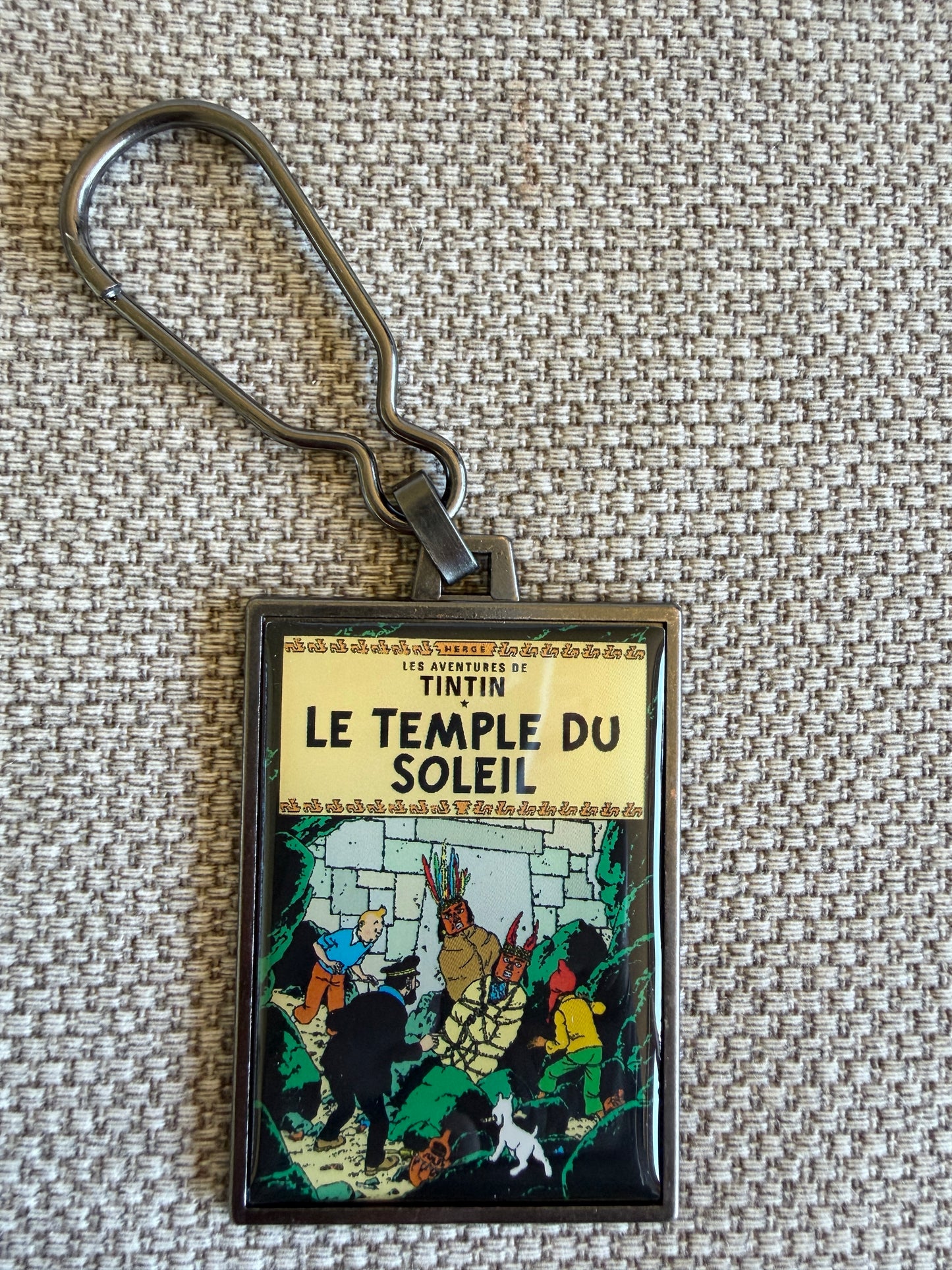 Front view of the rectangular Tintin Le Temple Du Soleil metal keychain on a woven fabric background, featuring a resin-coated replica of the original French book cover showing Tintin, Captain Haddock, Snowy, and Zorrino standing before the massive stone entrance of the Temple of the Sun.