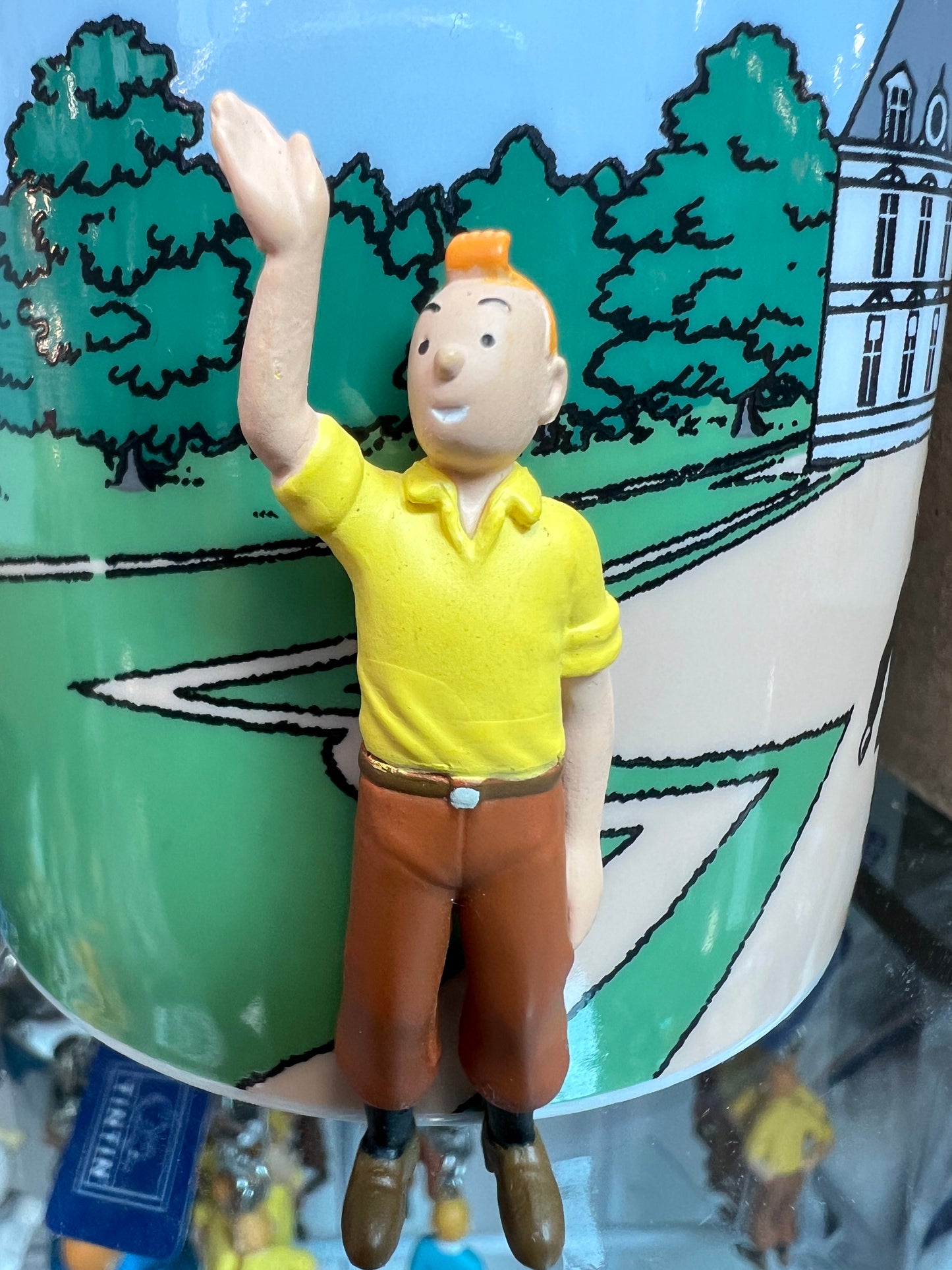 A close-up shot of the 6cm Tintin mini figure in a yellow polo shirt and brown trousers, raising his right hand in a wave, positioned against a mug featuring illustrated green trees and and Moulinsart castle.