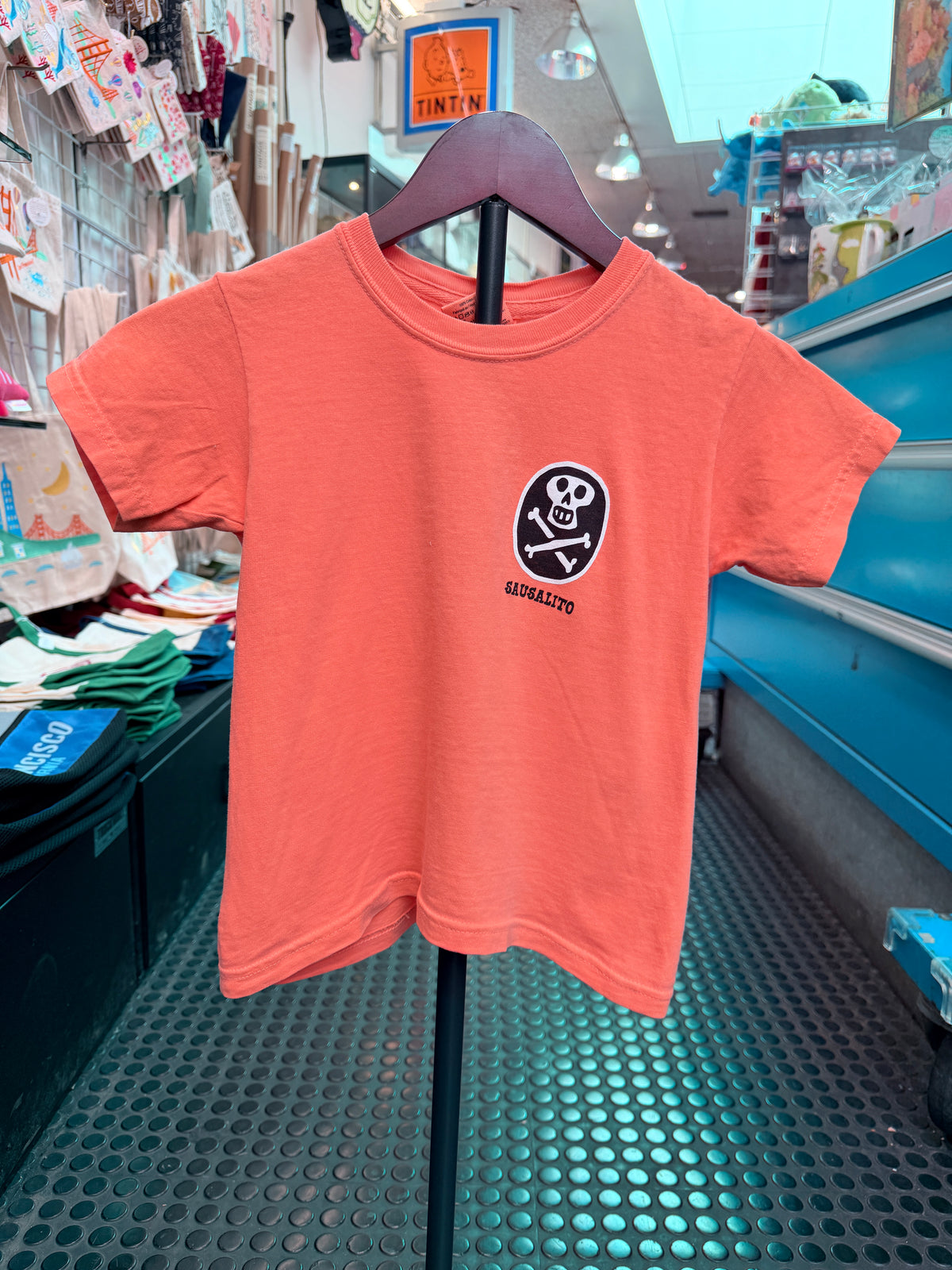 A melon-orange crewneck short-sleeve kid's t-shirt hanging on a display stand in a shop. The front chest features a small, black circular graphic containing a white skull and crossbones with the word 'SAUSALITO' printed directly underneath.