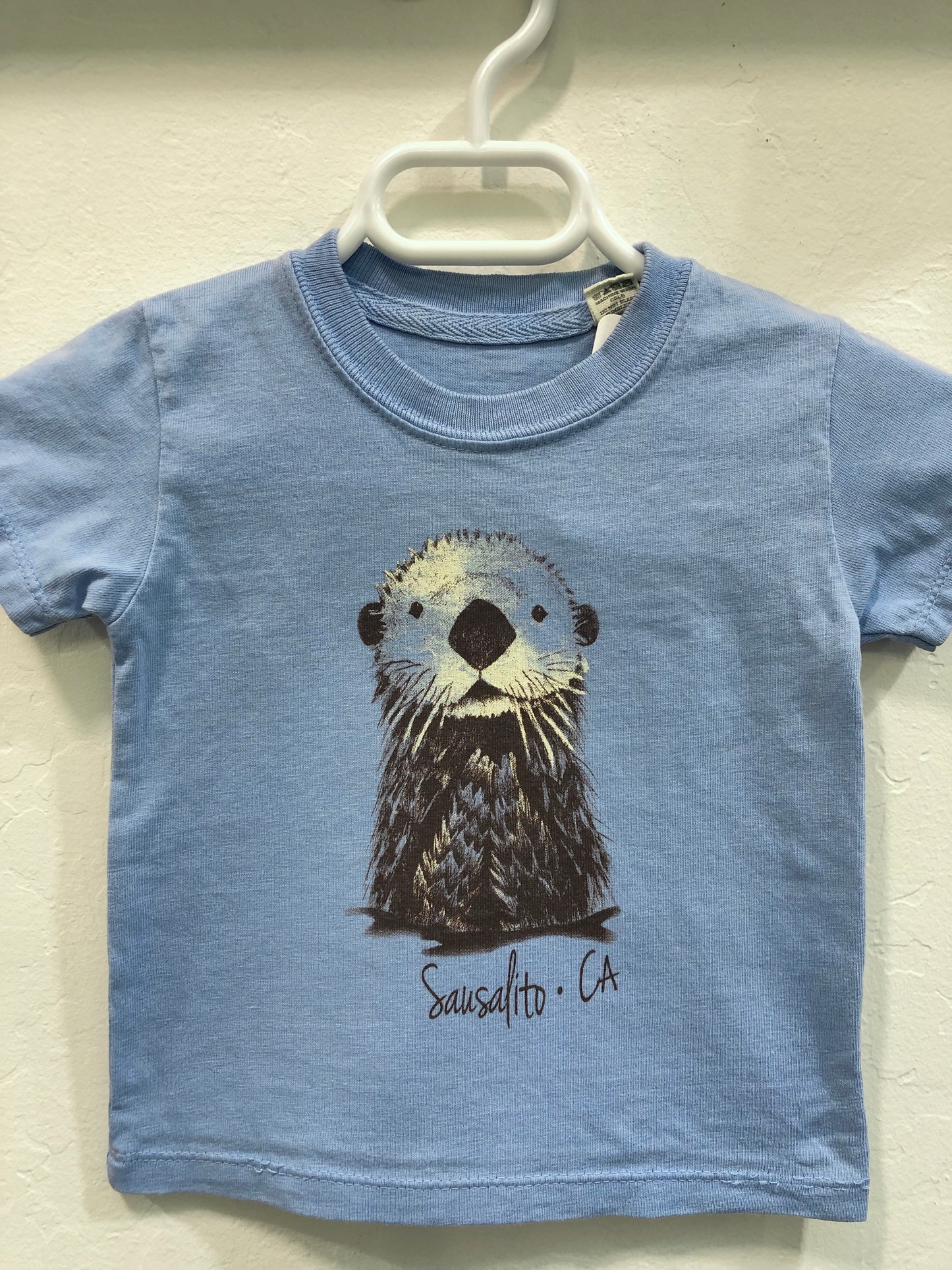 Blue infant t-shirt with cute sea otter illustration and 'Sausalito CA' text, hanging on white hanger