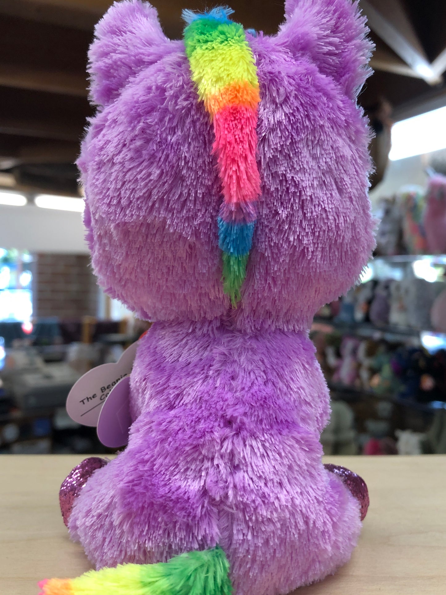 Ty Beanie Boo Rosette Unicorn plush toy with purple fur and rainbow mane and tail, shown from behind on store shelf