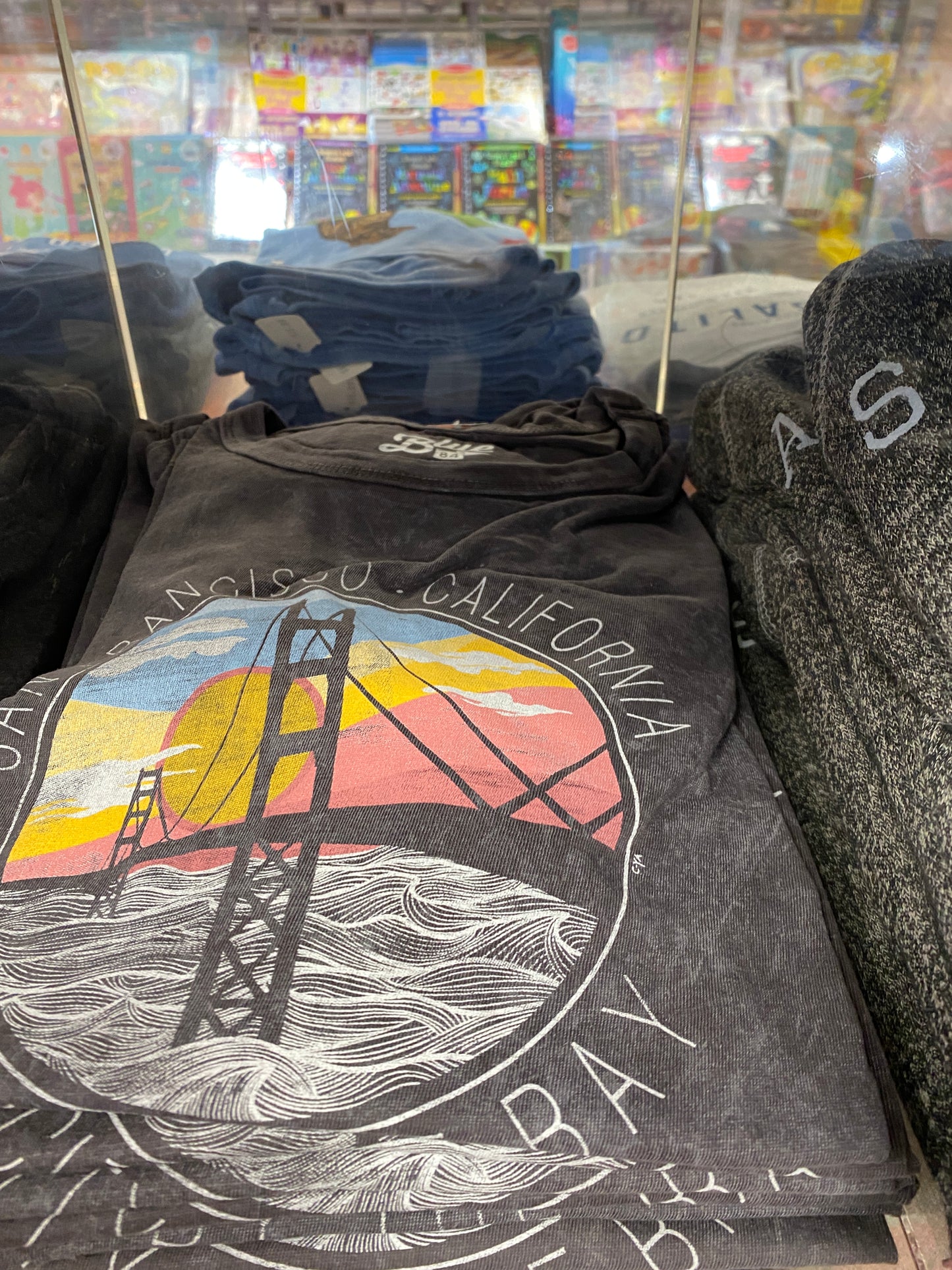 San Francisco Golden Gate Bridge t-shirt design with sunset and waves, displayed in souvenir shop alongside other clothing items