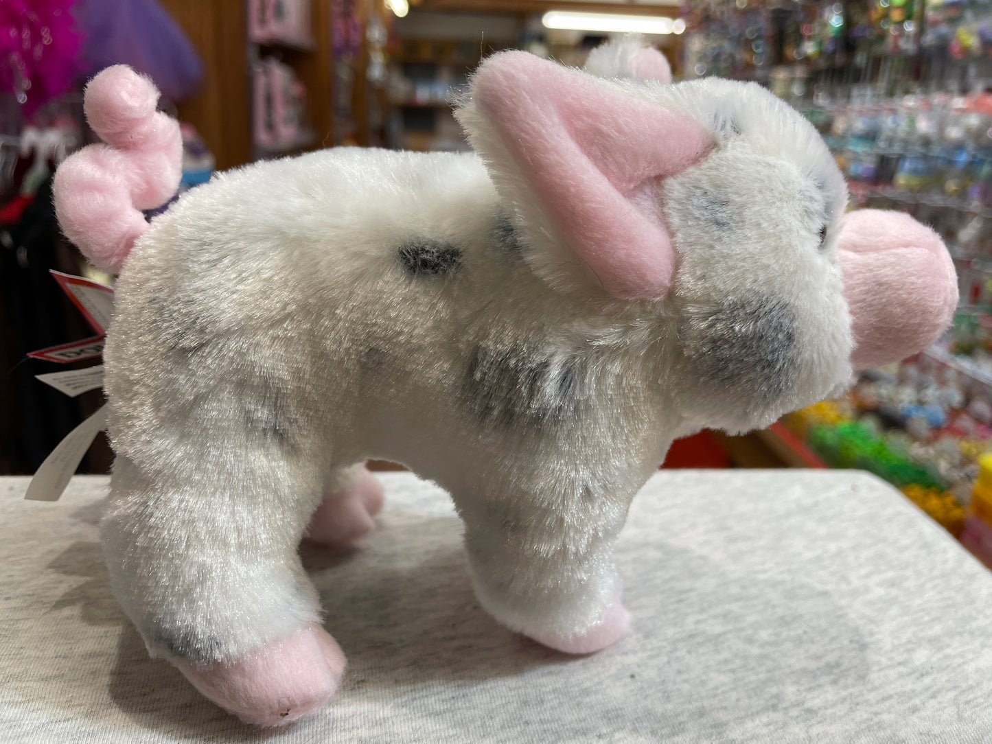 Soft plush black spotted pig toy with pink ears and curly tail, displayed in a colorful toy store setting