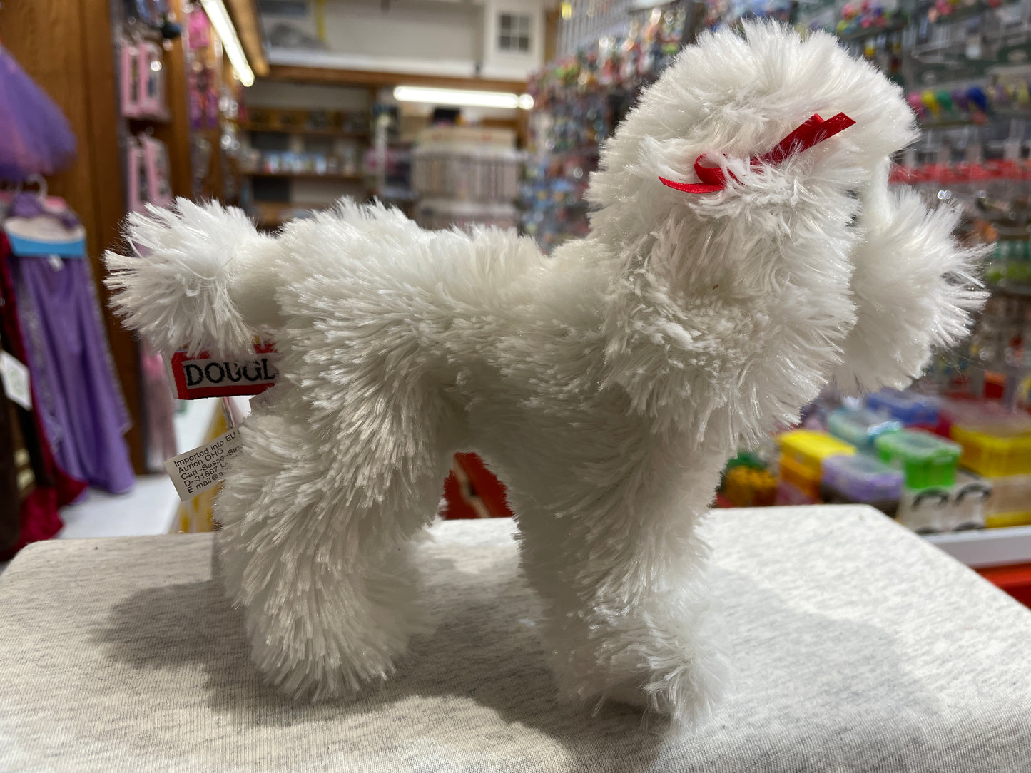 White poodle plush toy with red bow, standing on display in toy store, fluffy fur and classic poodle haircut visible