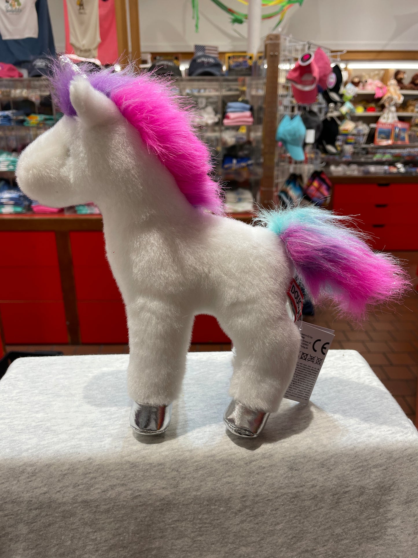 White unicorn plush toy with colorful pink and purple mane and tail, silver hooves, standing in store display