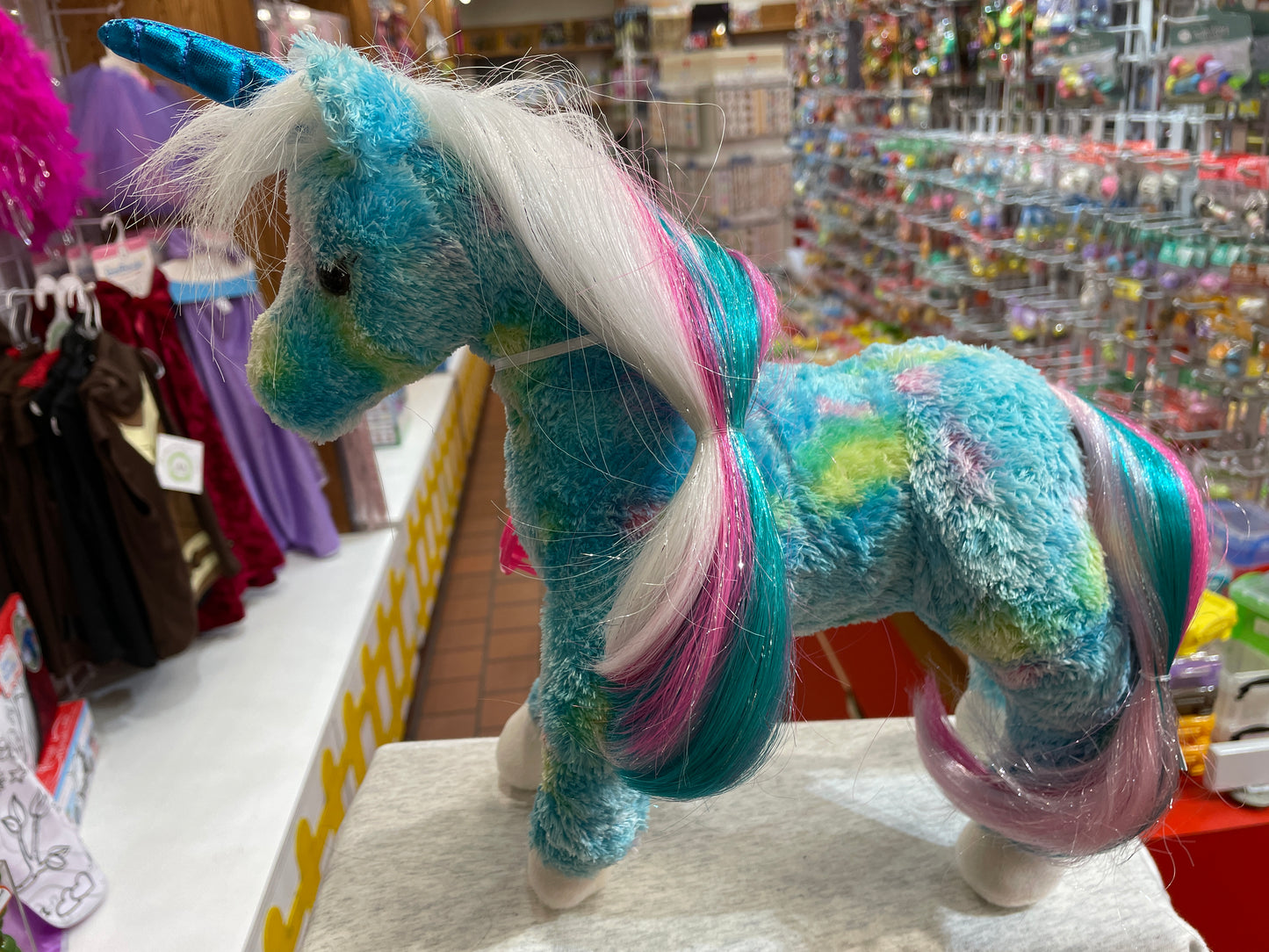 Colorful plush unicorn toy with blue coat and multicolored mane, displayed in toy store aisle with shimmering horn