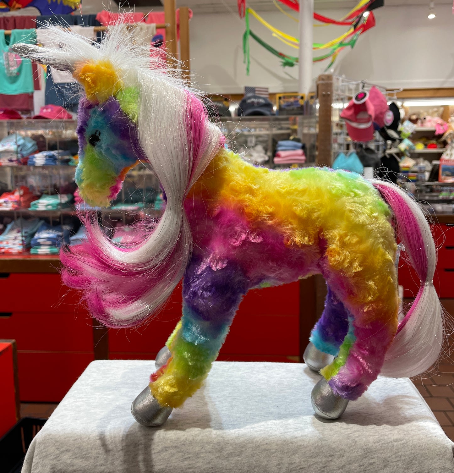 Rainbow unicorn plush toy with vibrant multicolored fur, long white and pink mane, silver hooves, and horn displayed in a store