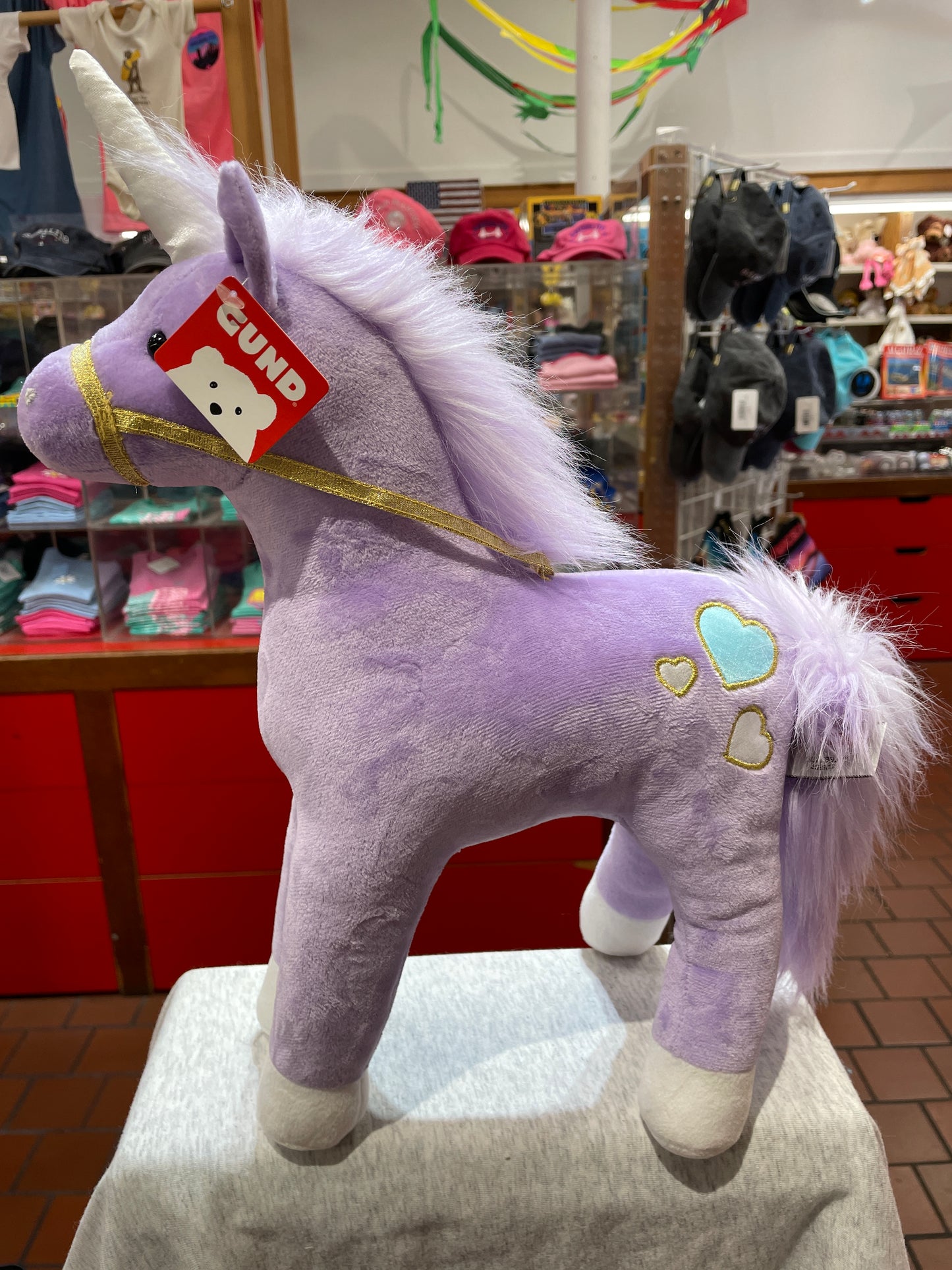 Gund Bluebell unicorn plush toy with purple fur, white mane, gold harness, and heart details in store display
