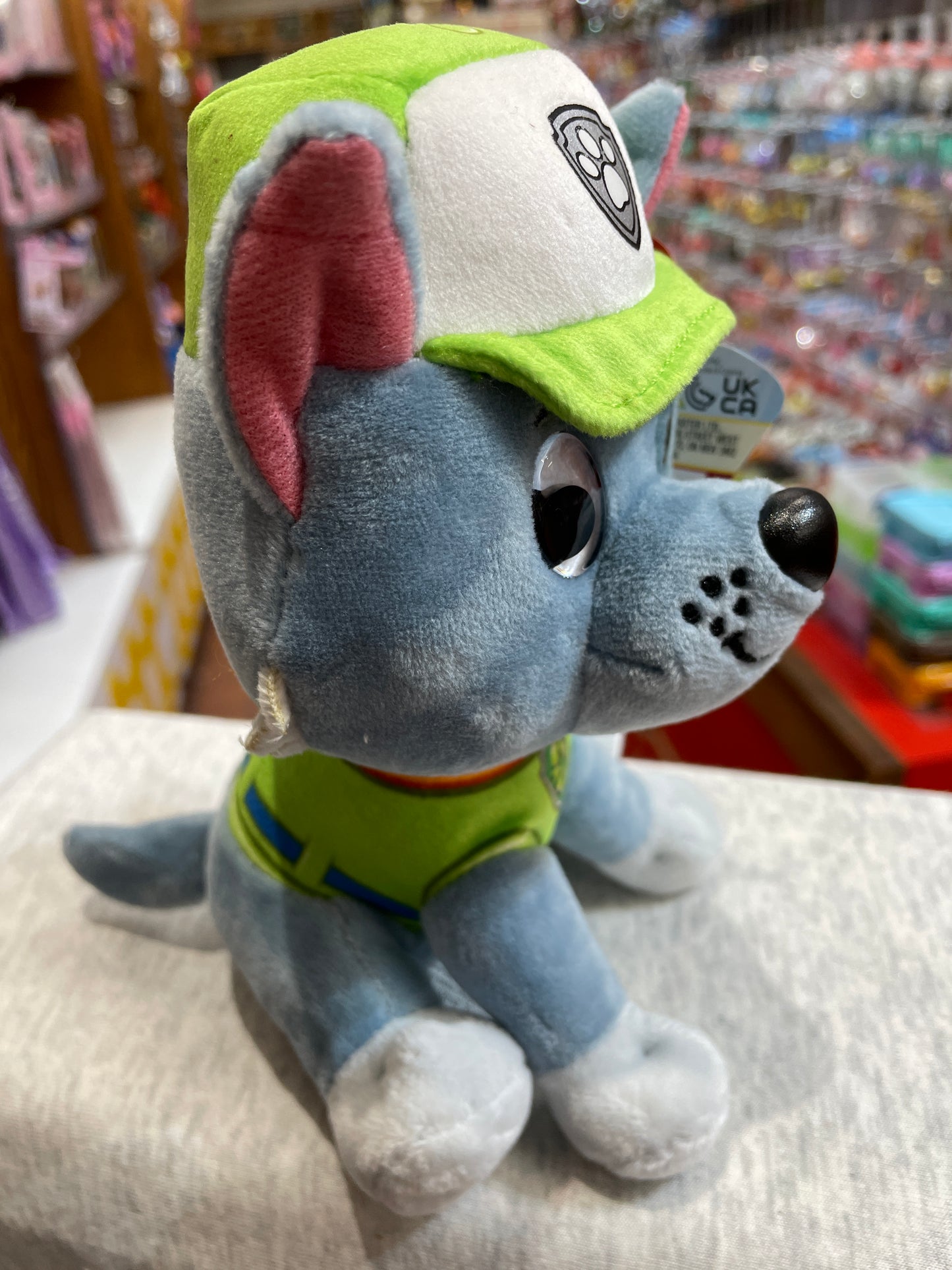 Gund Paw Patrol Rocky plush toy in recycling uniform with green cap, sitting on display in store