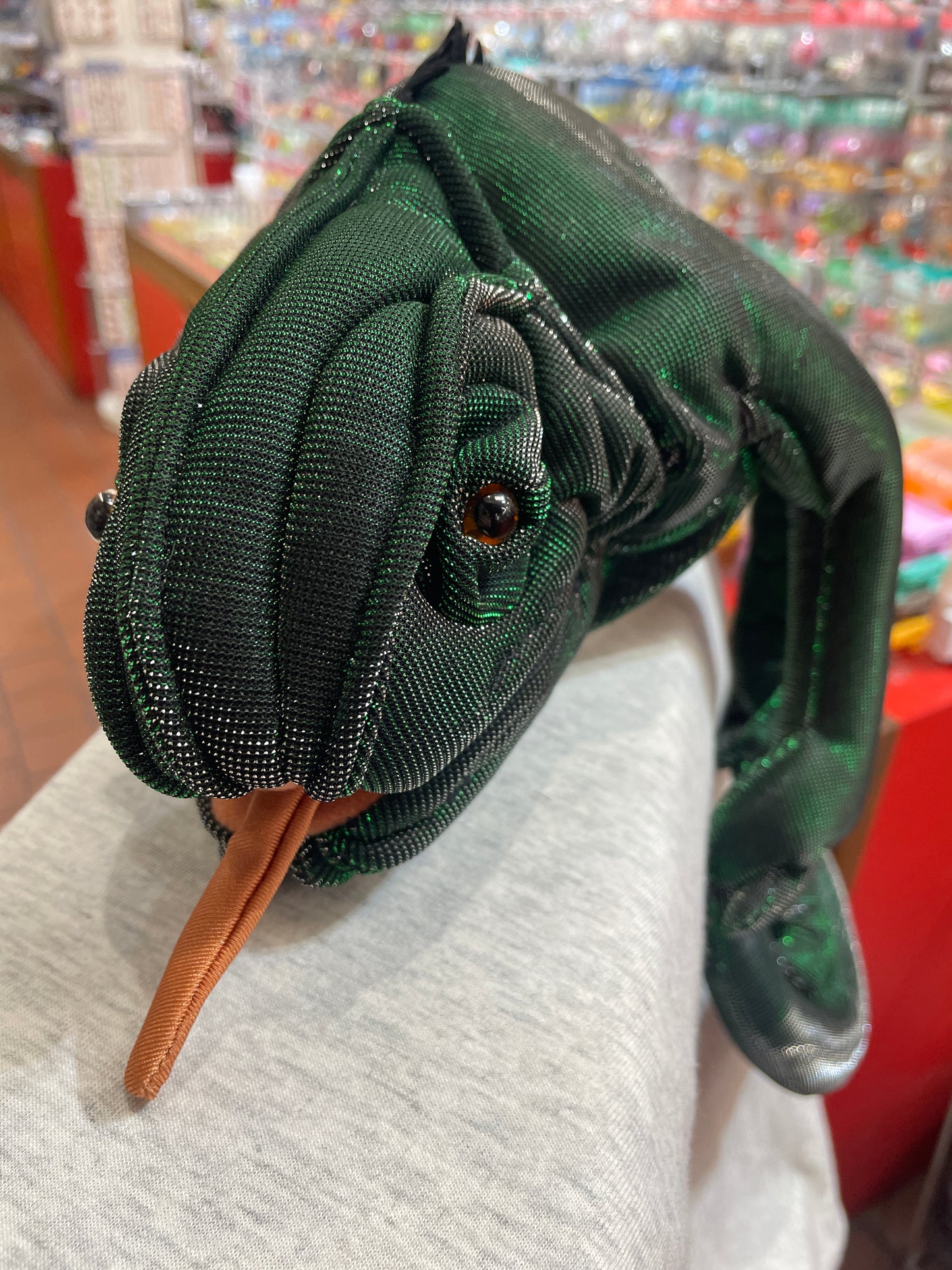 Shimmery green Folkmanis chameleon puppet with movable eyes and extended tongue, displayed in a toy store setting