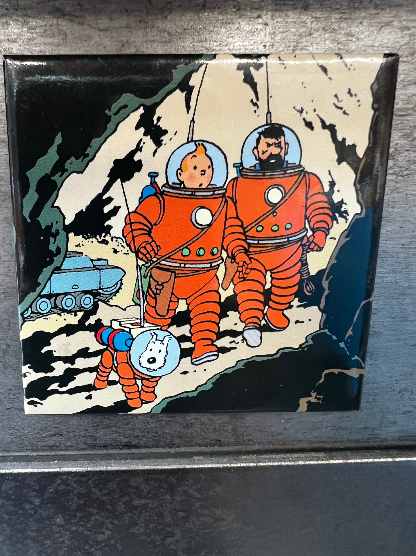Tintin moon magnet: Cartoon characters in orange spacesuits explore lunar cave entrance, with dog and vehicle nearby