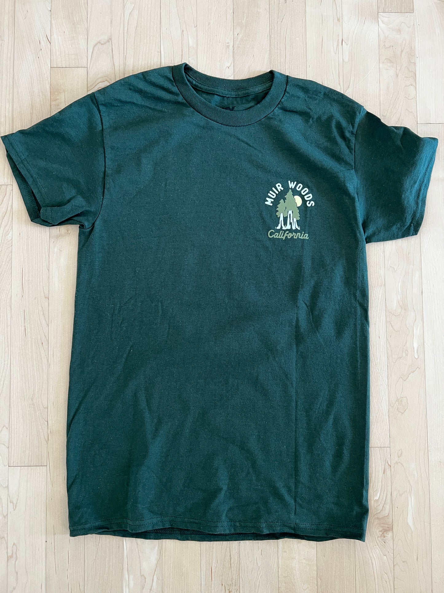 Green Muir Woods California t-shirt with redwood trees and sun logo on chest, displayed on wooden background