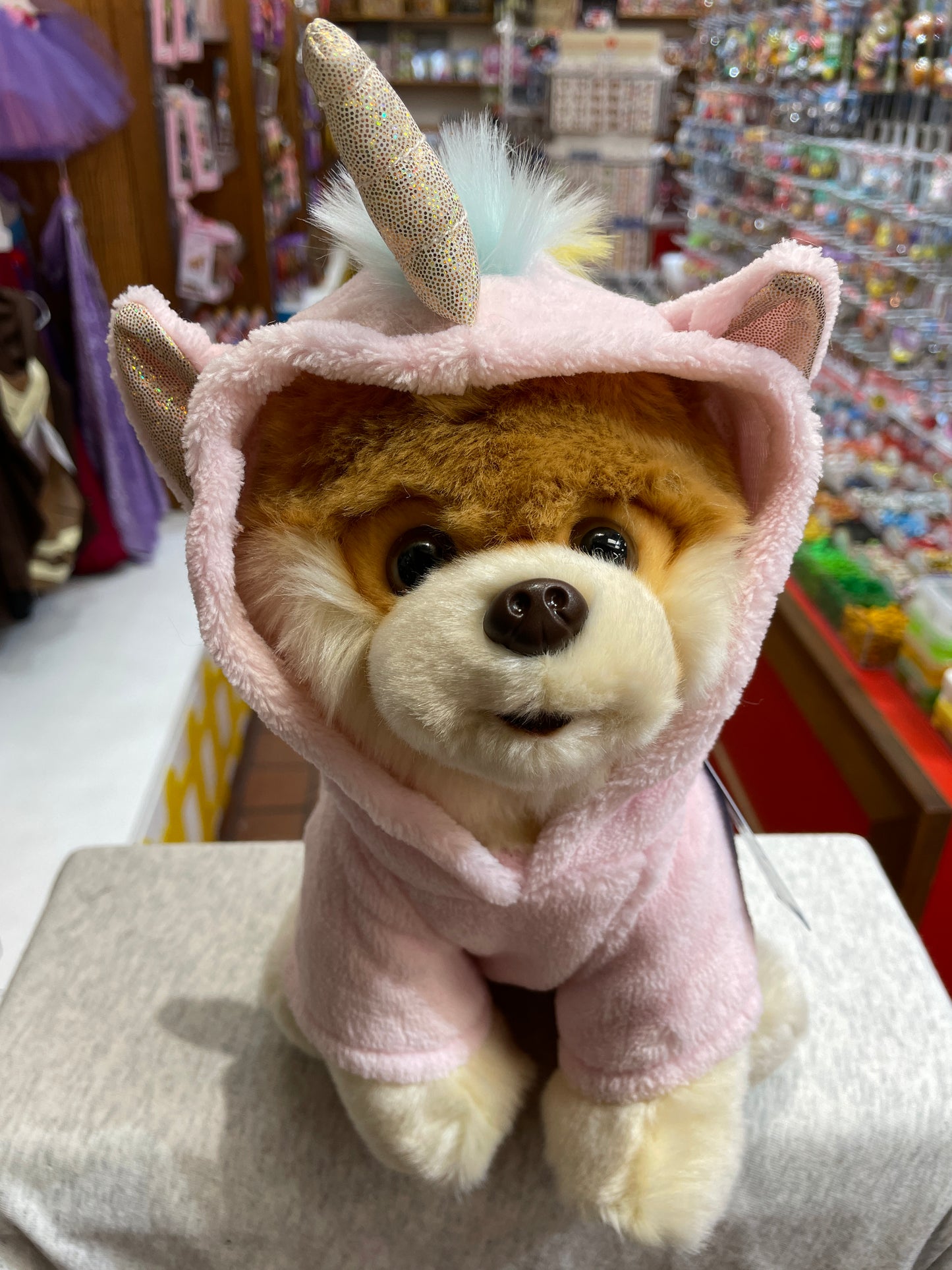 Gund Boo plush toy dressed in pink unicorn costume with sparkly horn and ears, displayed in toy store