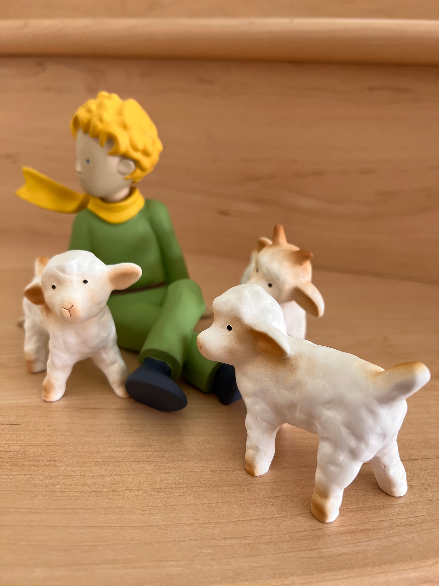 Le Petit Prince resin figures: blonde character in green outfit sitting with three white sheep figurines on wooden surface