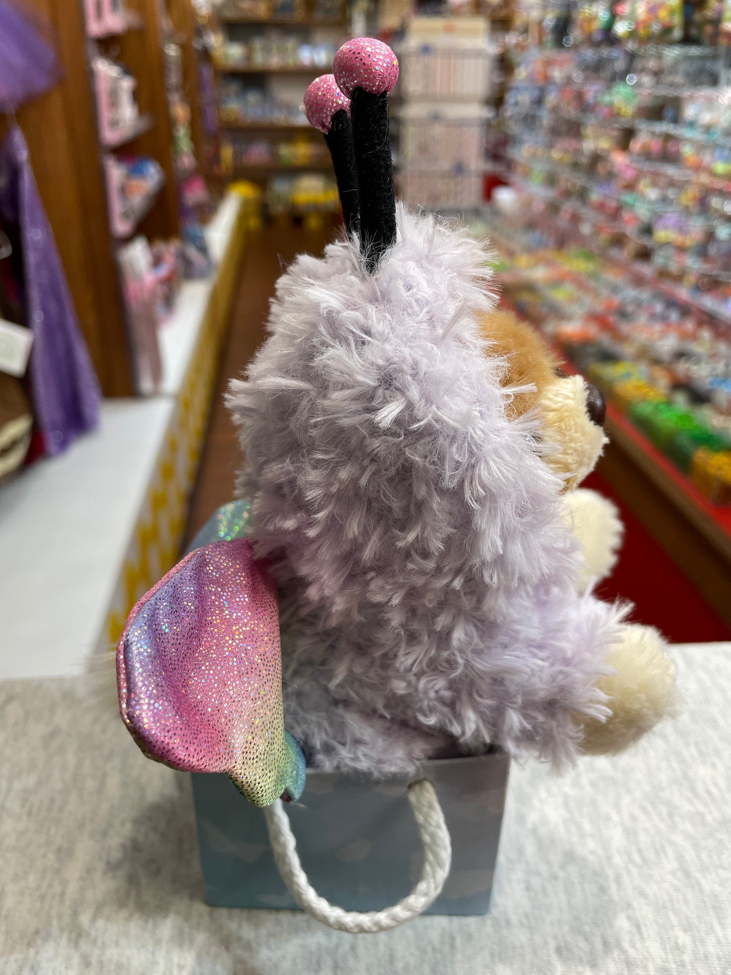 Gund Boo plush toy in butterfly costume with shimmering wings, peeking out of gift bag in toy store aisle