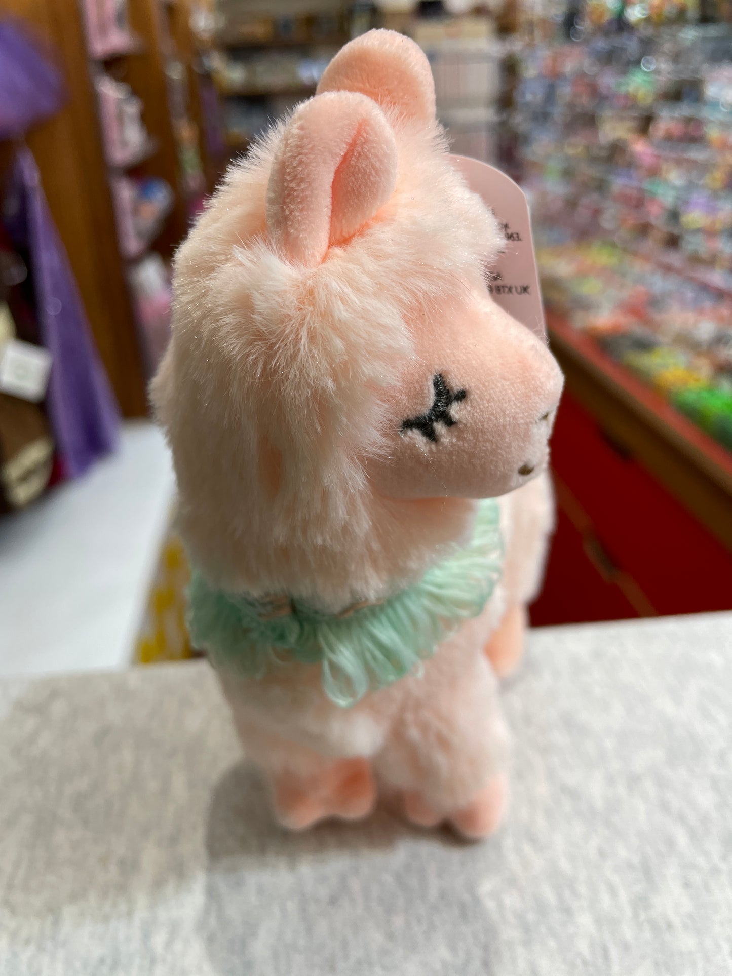 Soft pink Mary Meyer Lexi Llama plush toy with light teal neck scarf, displayed in toy store setting