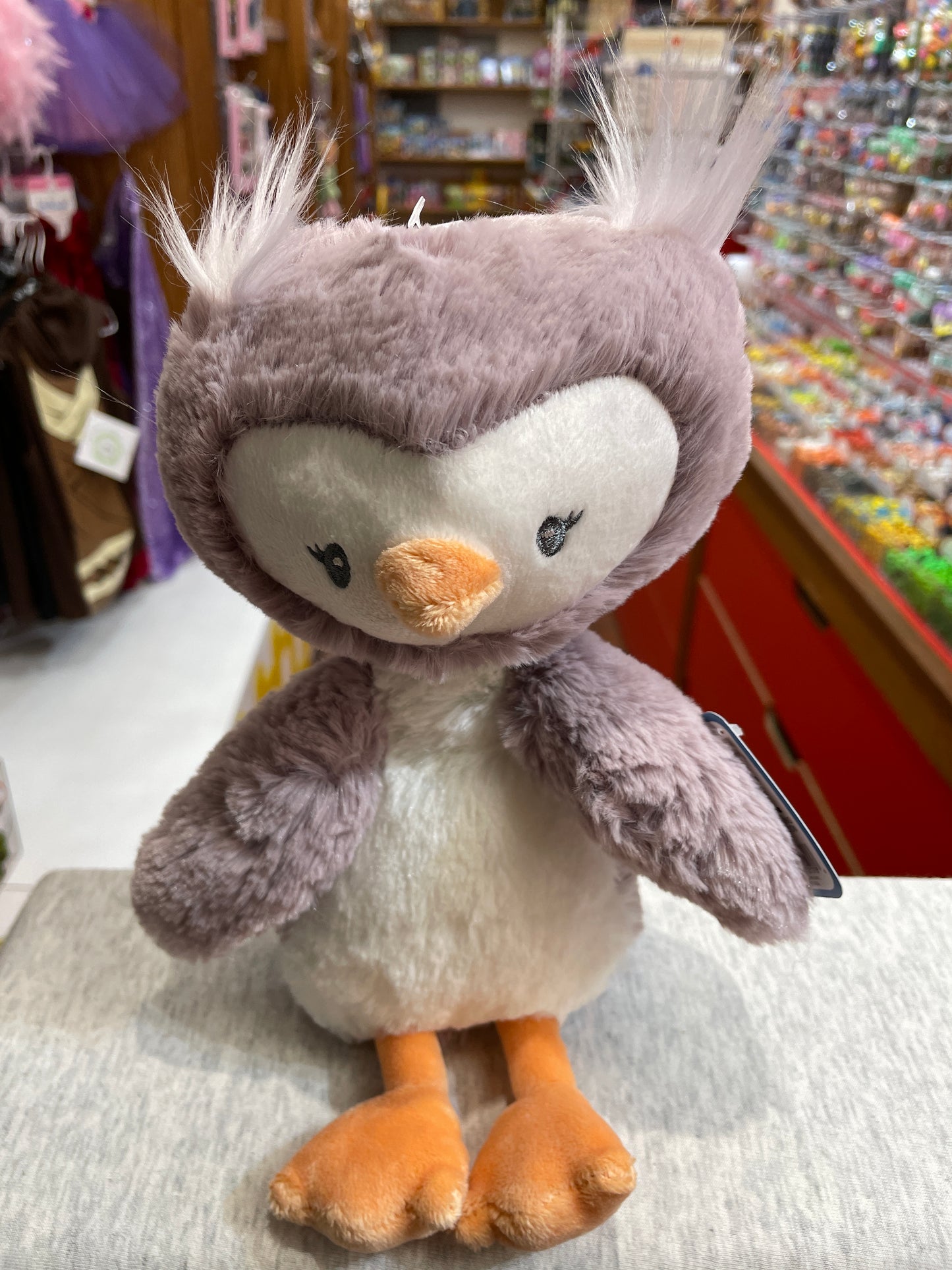 Soft purple Baby Gund Toothpick Owl plush toy with fuzzy feathers, embroidered eyes, and orange feet in toy store setting