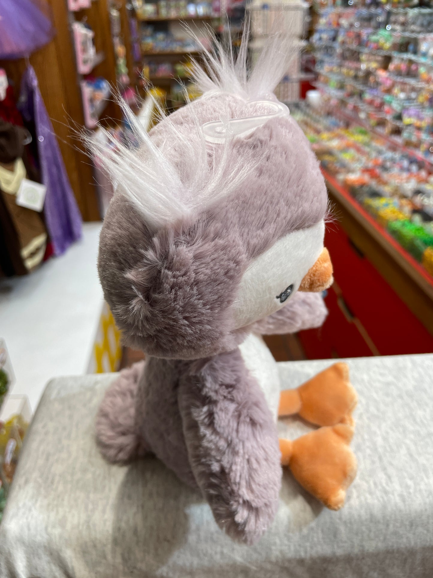 Soft purple Baby Gund Toothpick Owl plush toy with fuzzy white feathers and orange feet, displayed in a toy store