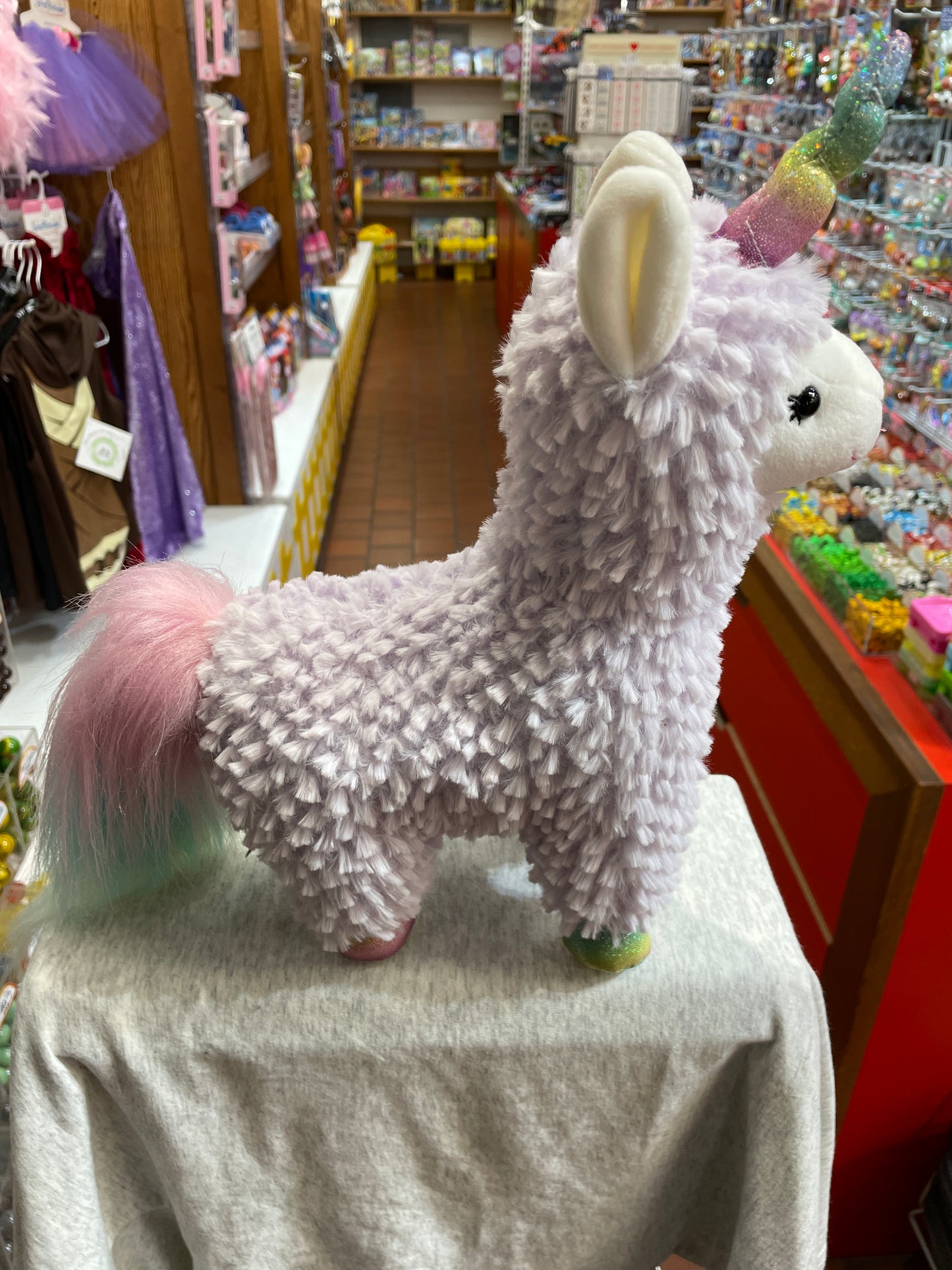 Gund Sugar Plum Llamacorn plush toy with sparkly unicorn horn and rainbow tail, displayed in colorful toy store aisle