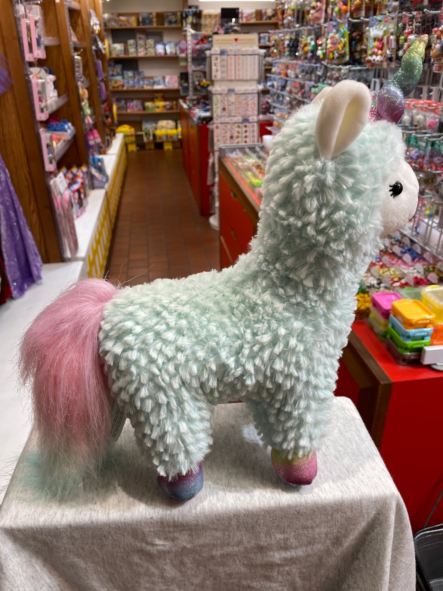 Gund Cotton Candy Llamacorn plush toy in pastel turquoise with pink tail and rainbow hooves, displayed in a toy store aisle
