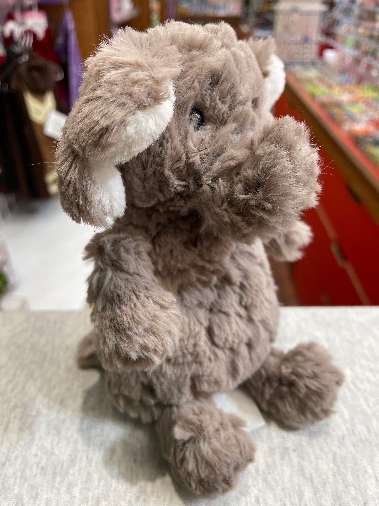 Soft, fluffy gray Mary Meyer Puttling Elephant plush toy sitting on display, featuring large floppy ears and a sweet smile