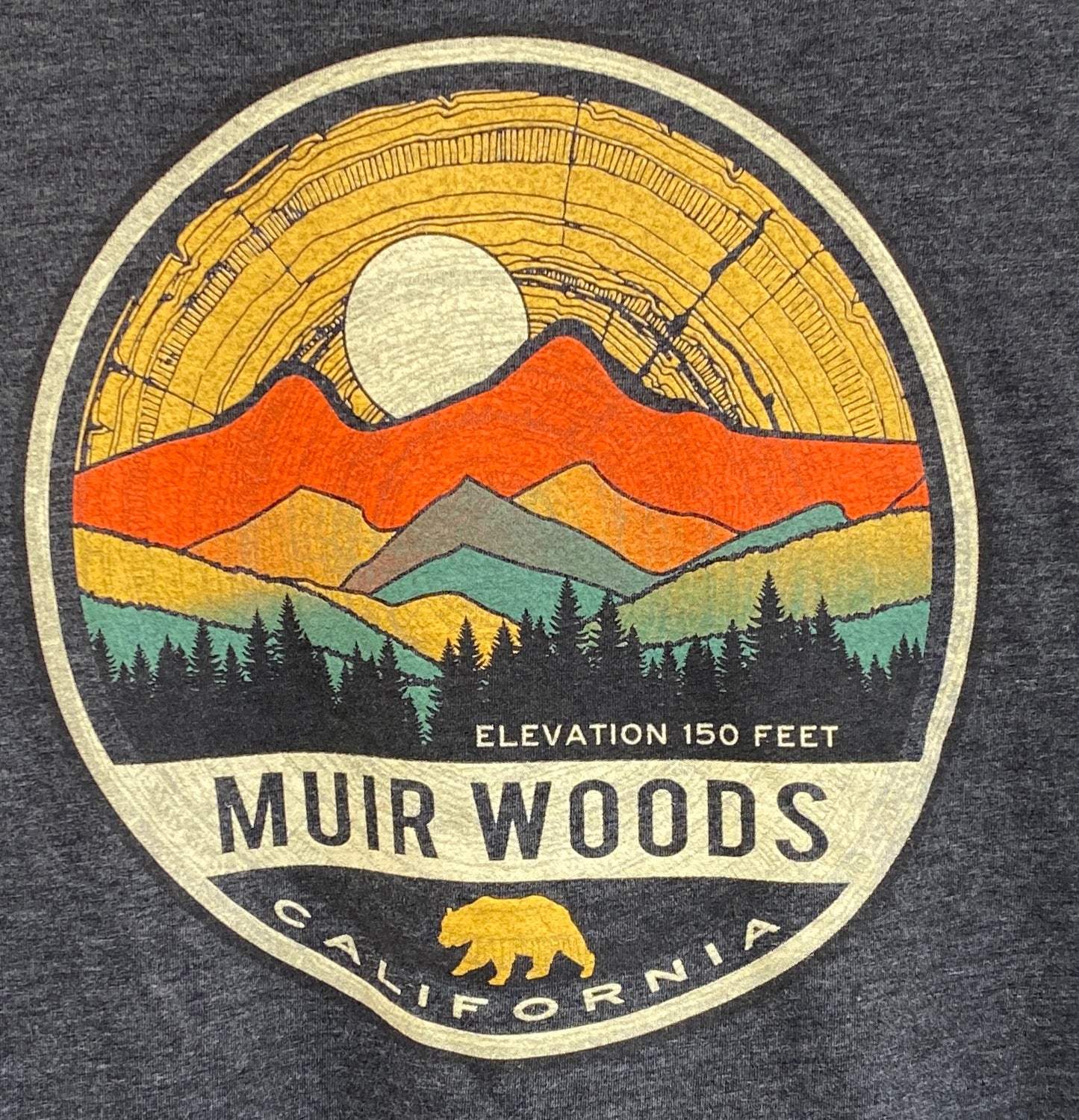 Muir Woods t-shirt design featuring colorful mountain landscape, sunset, and forest silhouette in circular emblem with California bear logo