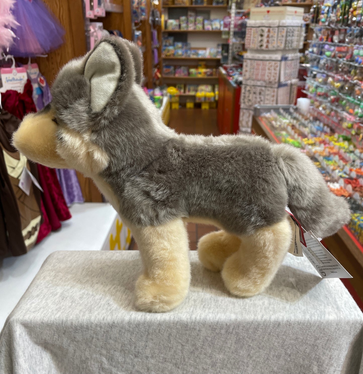 Soft gray and tan wolf plush toy standing alert on display in toy store, surrounded by colorful merchandise
