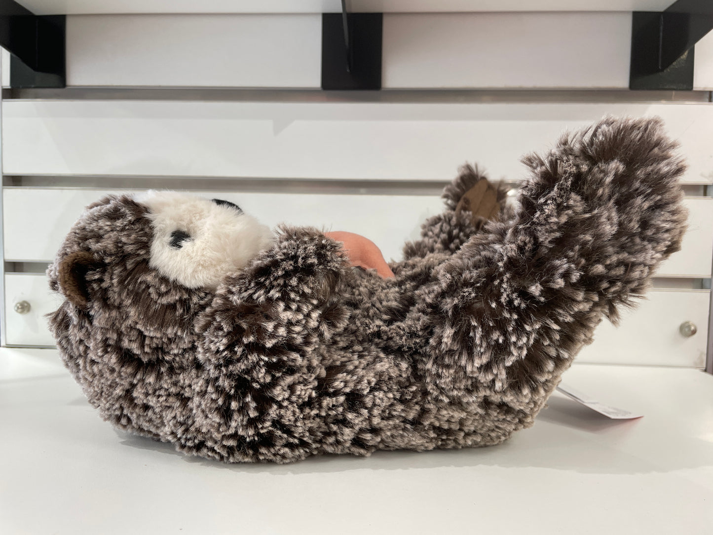 Cuddly sea otter plush toy with realistic gray-brown fur, white face, and curved body, lying on its back on a white surface