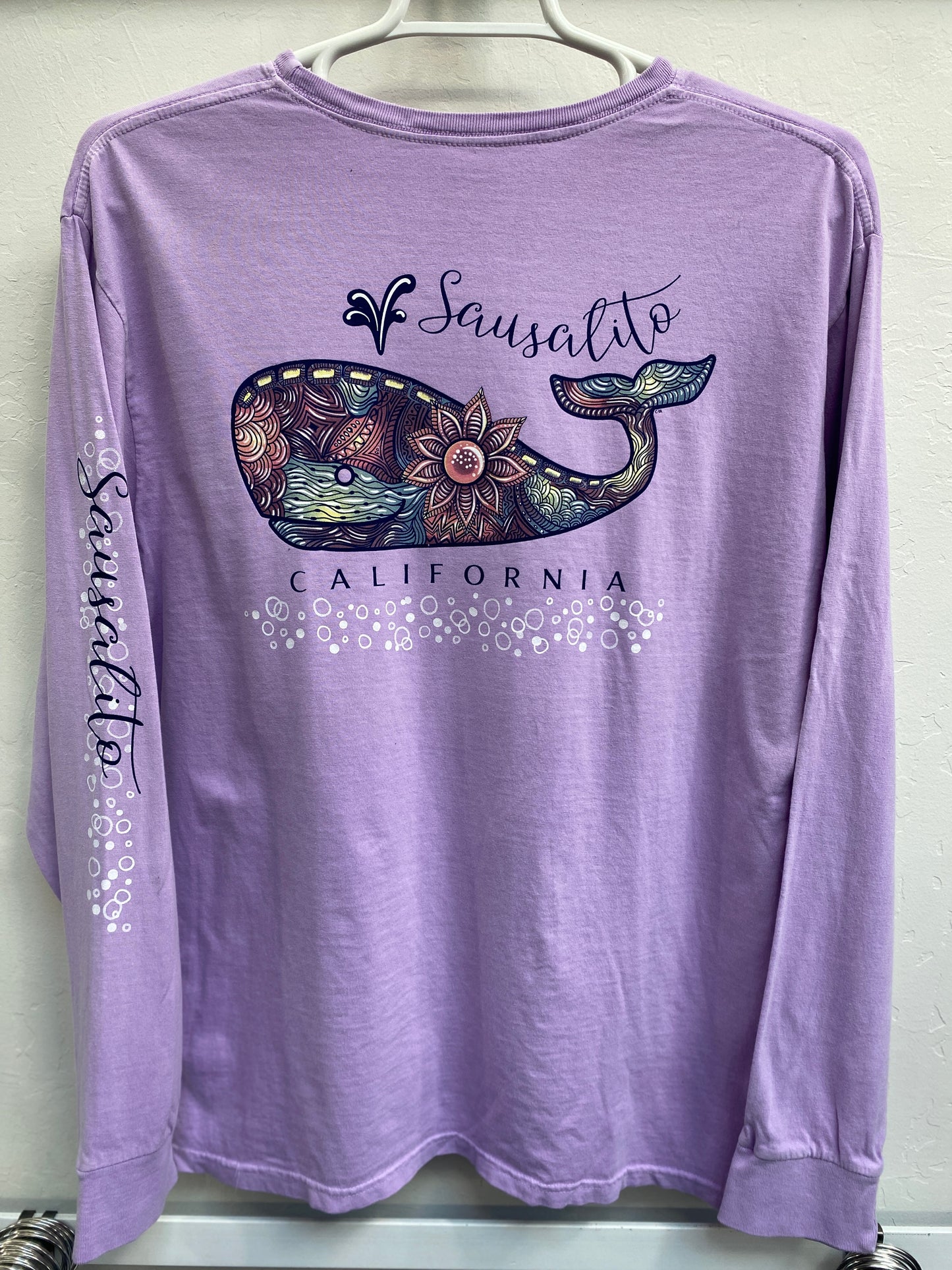 Lavender long sleeve shirt with colorful mandala whale design and Sausalito California text, hanging on display