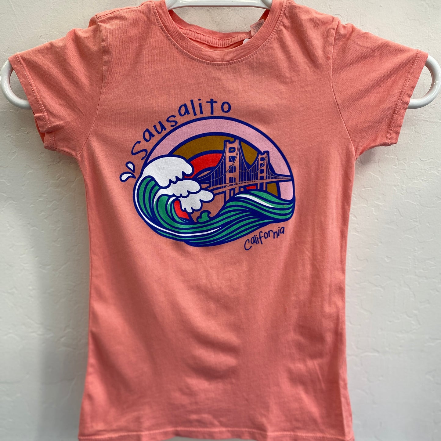 Coral kids' t-shirt with cartoon Sausalito design featuring Golden Gate Bridge and wave against sunset, California souvenir