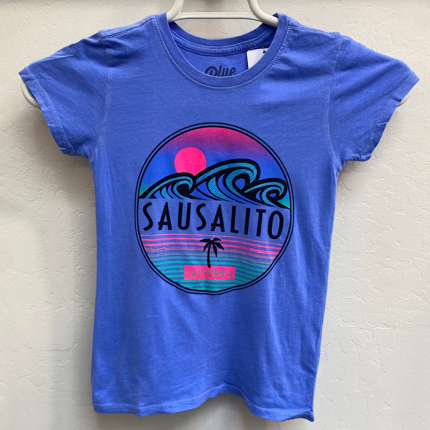 Blue girls' t-shirt with retro Sausalito California graphic featuring sunset, ocean waves, and palm tree in vibrant colors