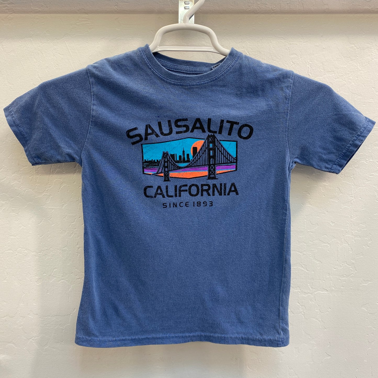 Blue kids' t-shirt with colorful Golden Gate Bridge design, Sausalito California Since 1893 text, hanging on white hanger