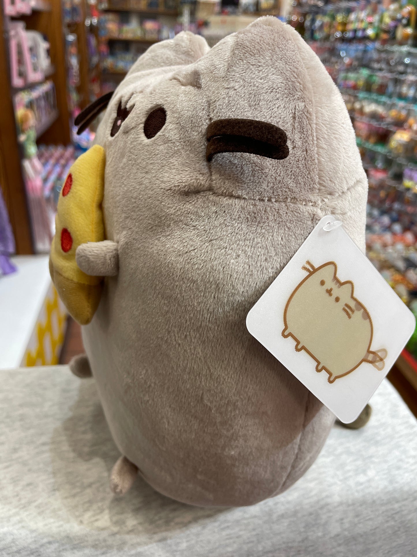Gund Pusheen plush toy holding pizza slice, displayed in store with character tag, soft gray tabby cat design