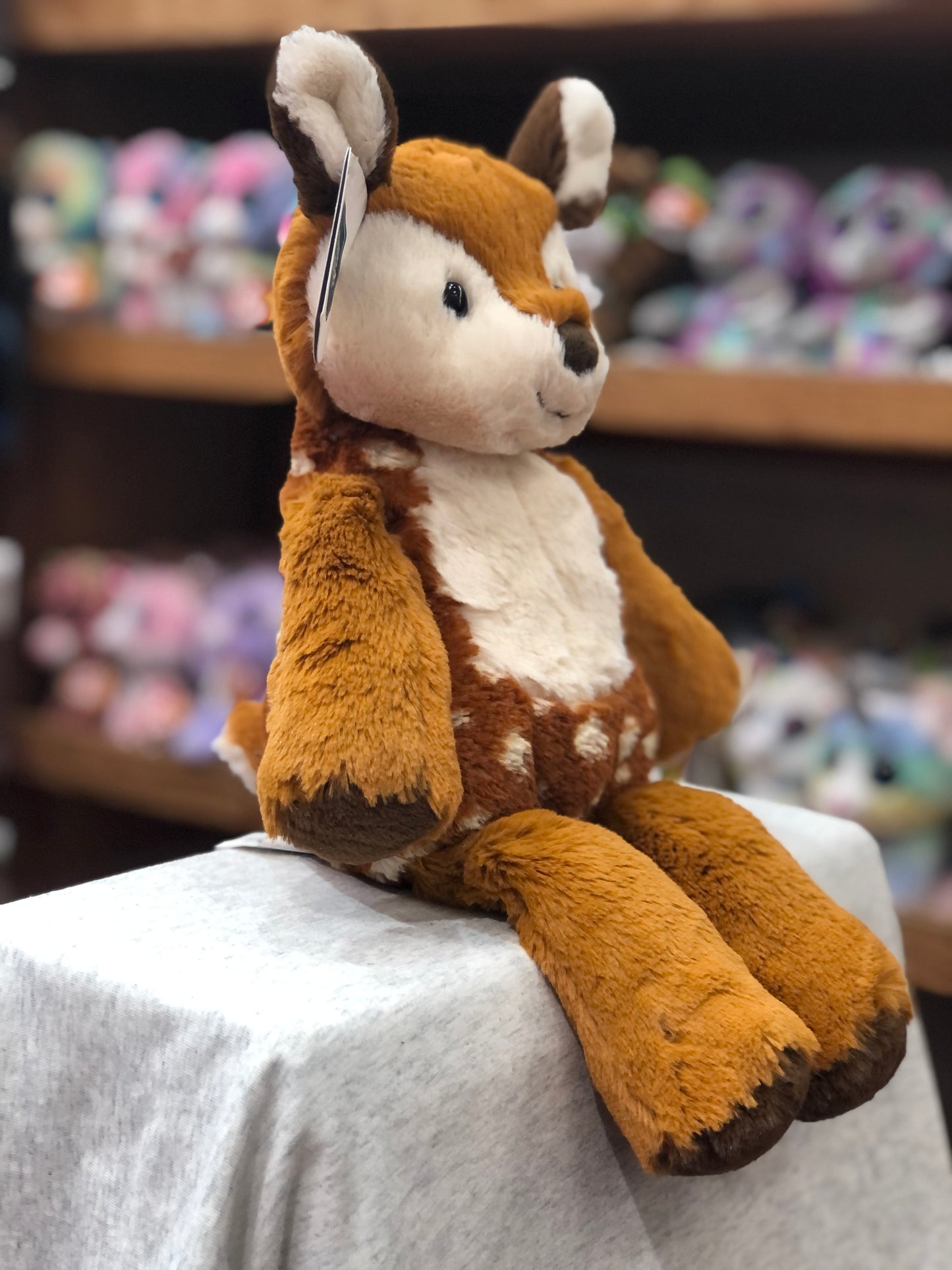 Soft and cuddly Mary Meyer Marshmallow Fawn plush toy sitting on display, with colorful stuffed animals in background