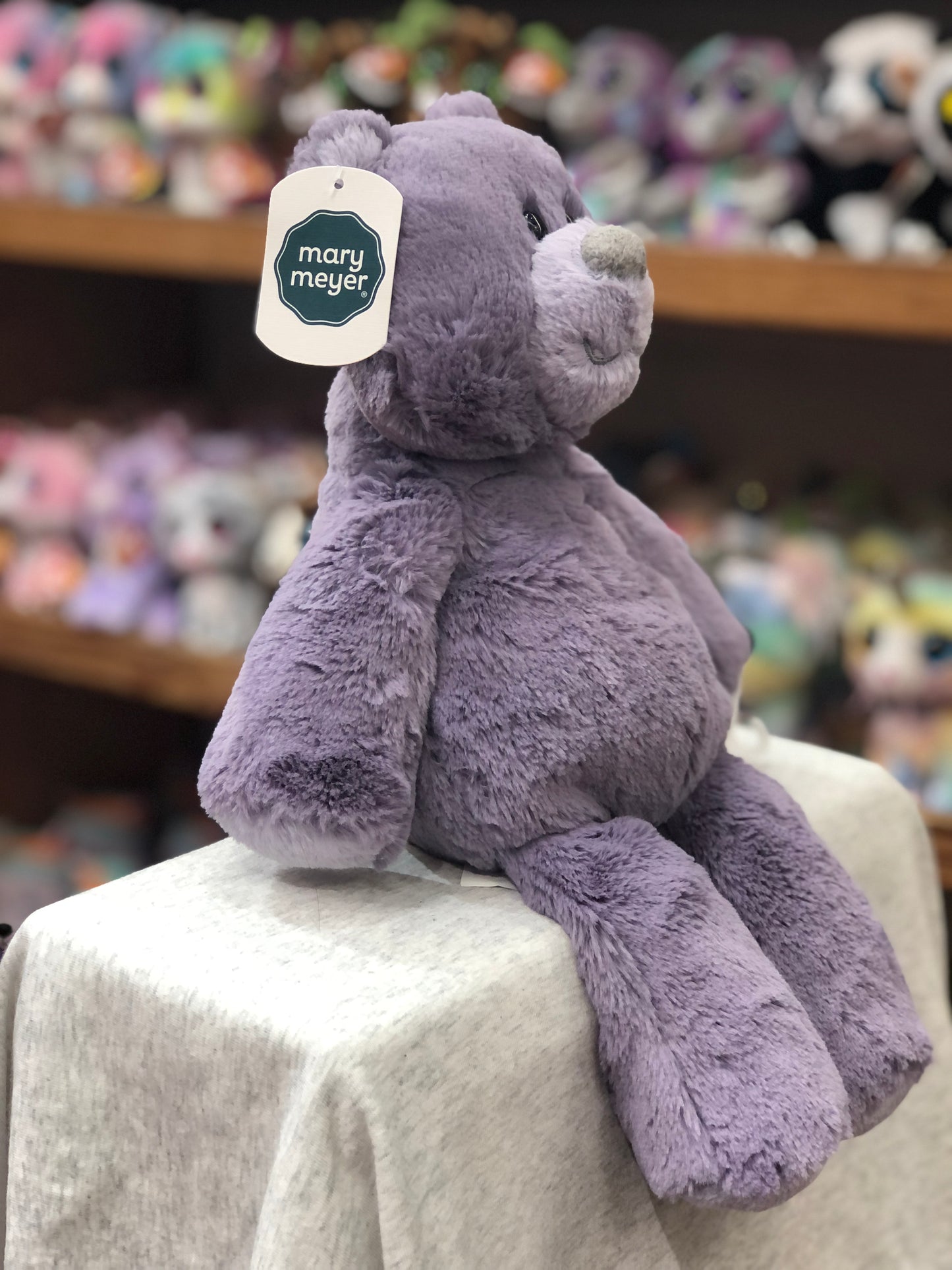 Soft purple Mary Meyer Marshmallow Berry Bear plush toy sitting on display, with tag visible and colorful stuffed animals in background