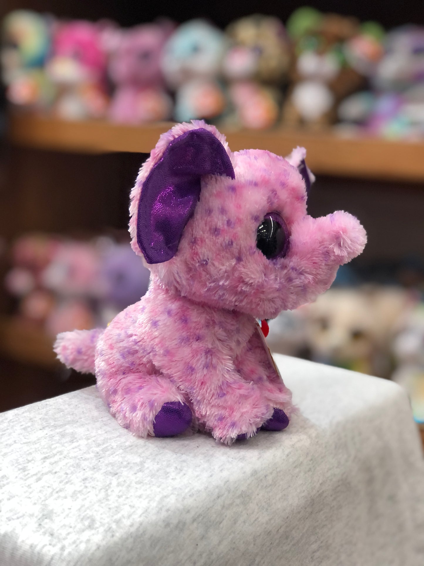 Pink speckled Ty Beanie Boo elephant plush with purple ears, sitting on display with other plush toys in background