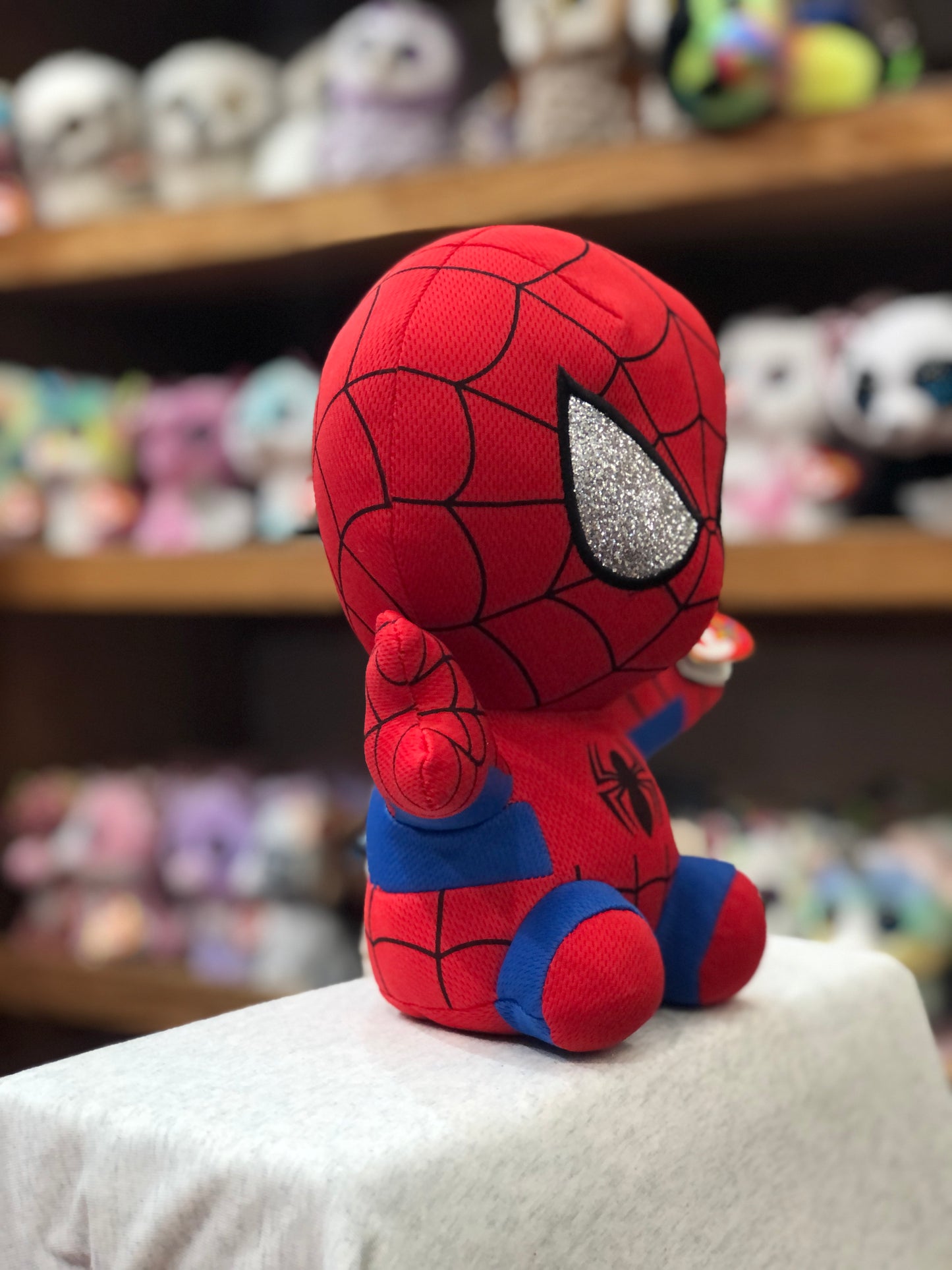 Ty Beanie Buddy Spiderman plush toy with glittery eye, displayed on shelf with other plush toys in background