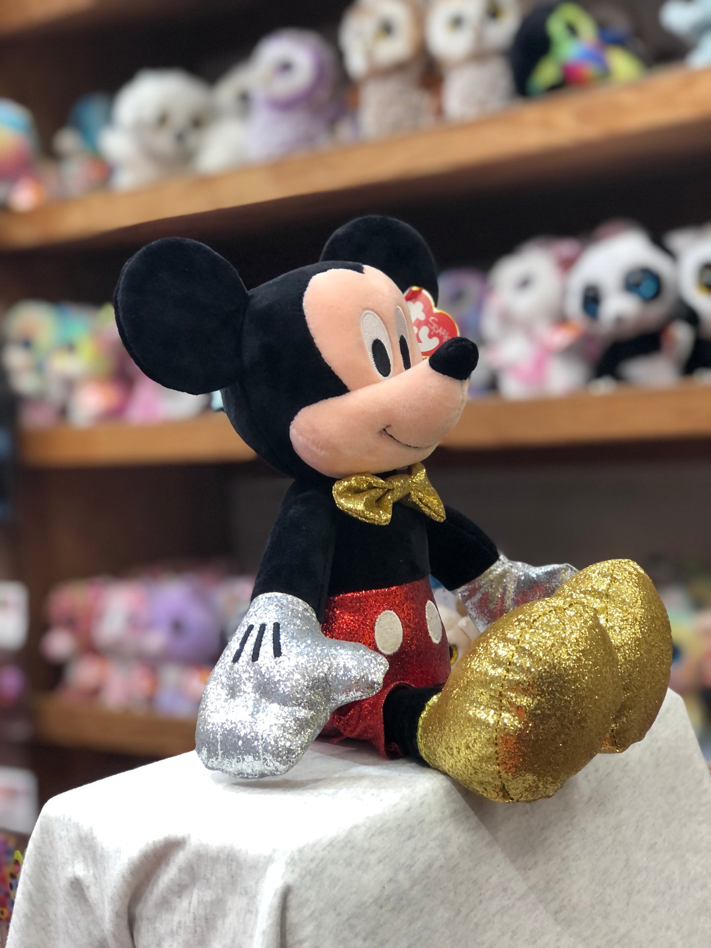 Mickey Mouse plush toy with sparkly red shorts, gold bow tie, and glittery silver gloves on display with other plush toys