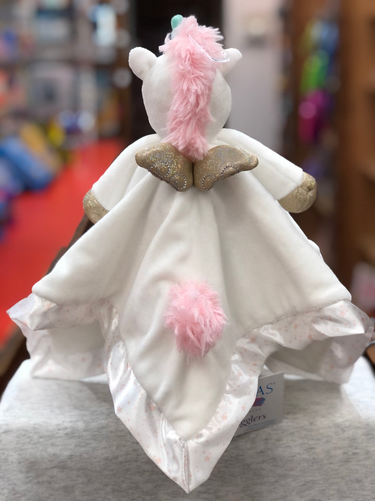 Soft white unicorn plush toy with pink mane, teal horn, and golden wings. Features star-speckled border and pink tail on blanket body.