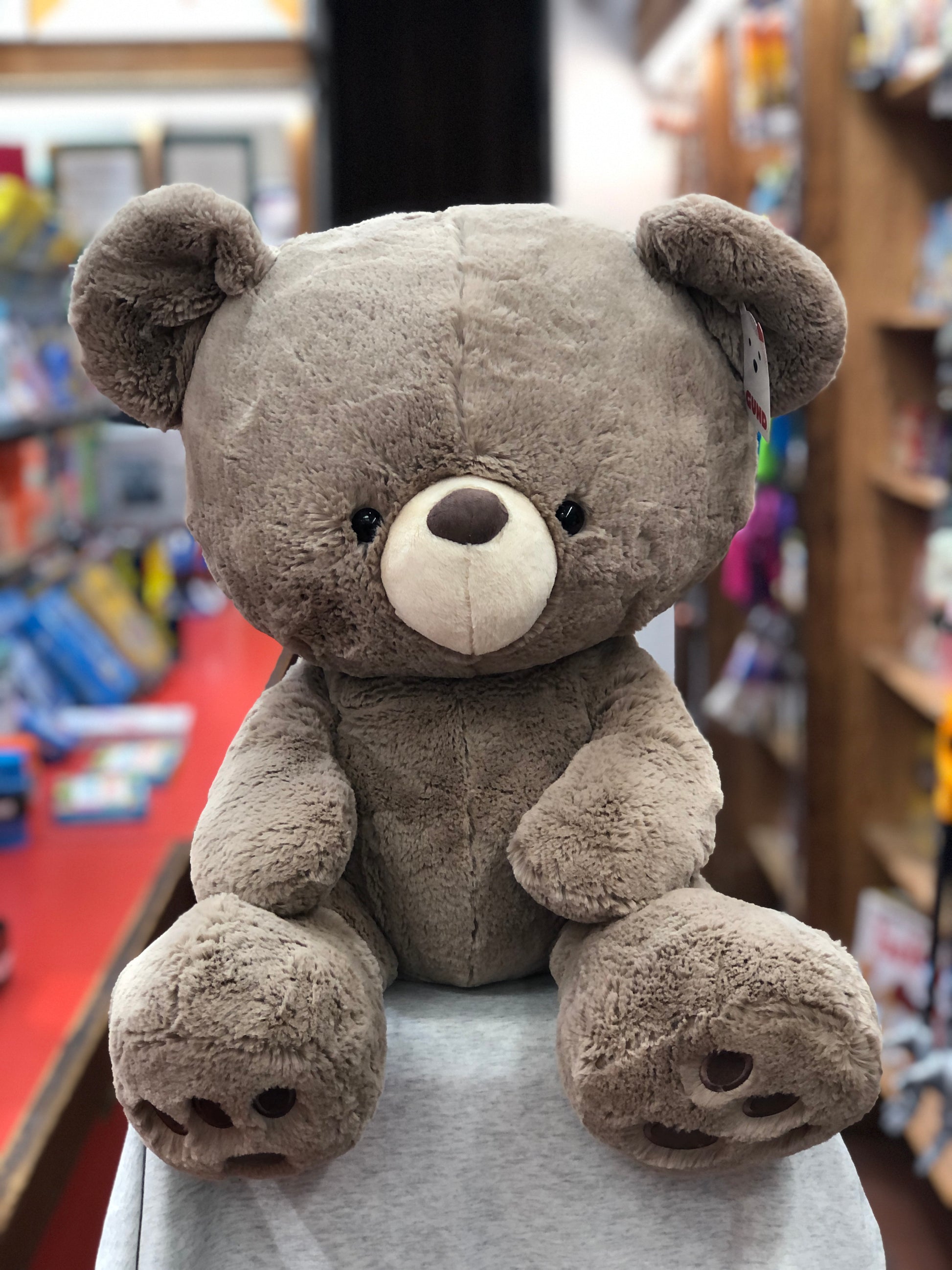 Large 23-inch Gund Kai Bear plush toy in taupe, with oversized head and embroidered paw pads, sitting on display in toy store