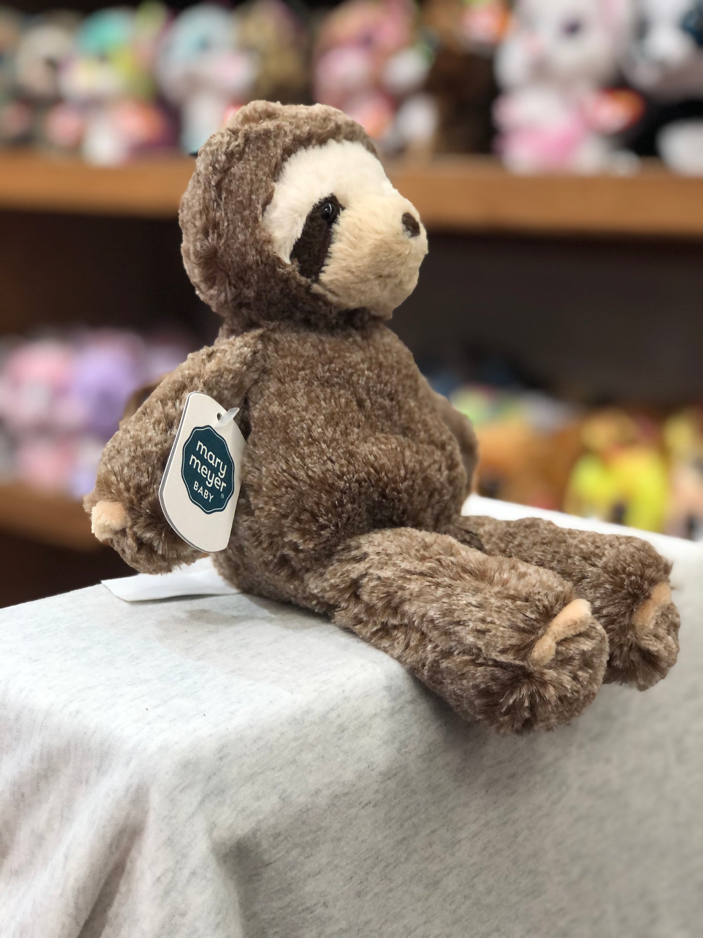 Soft, cuddly Mary Meyer Marshmallow Zoo Junior Sloth plush toy sitting on display, with colorful stuffed animals in background