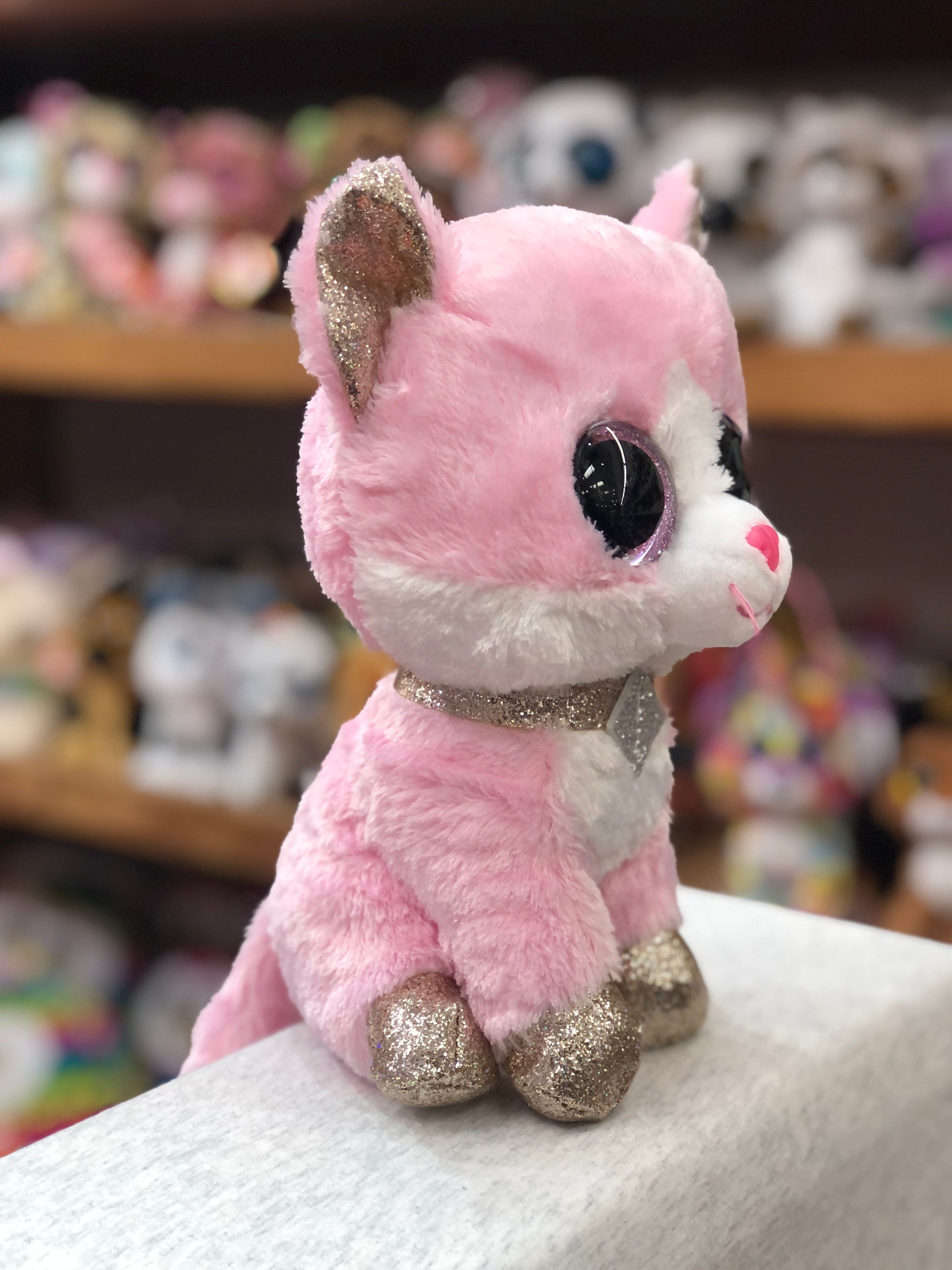 Ty Beanie Boo pink cat plush Fiona with glittery ears, feet, and sparkly necklace, sitting among other stuffed toys