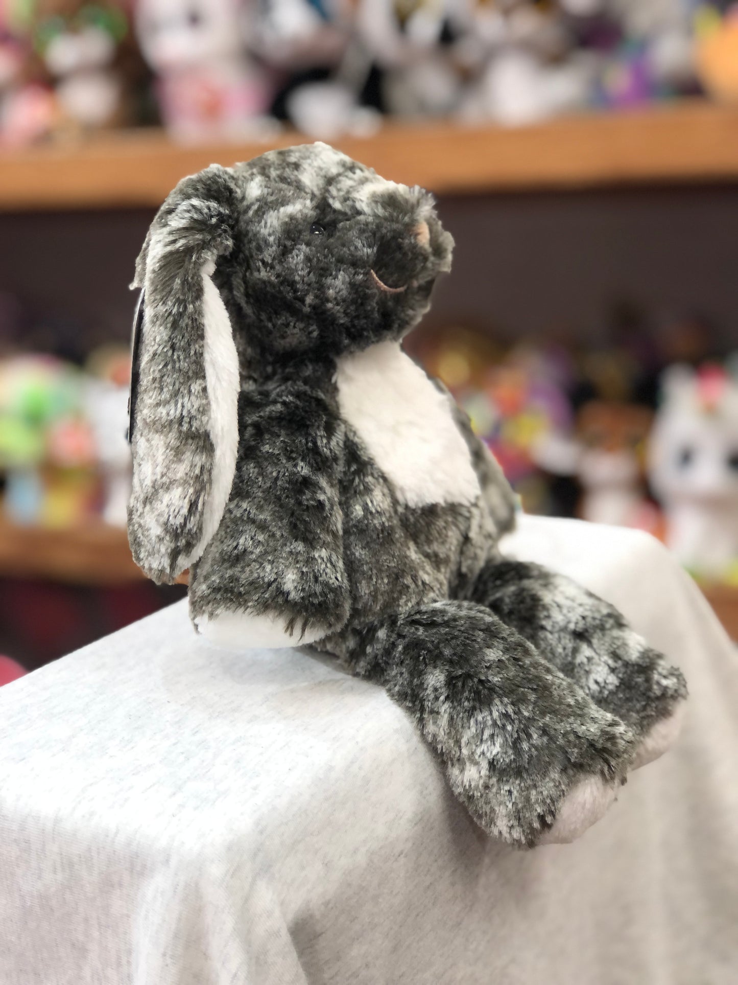 Soft gray plush bunny from Marshmallow Zoo Junior collection, sitting on white surface with colorful toys in background