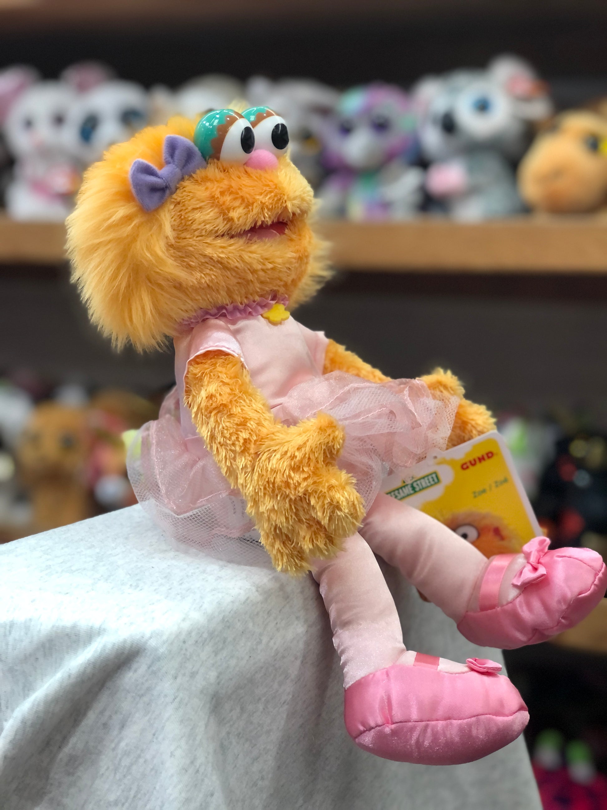 Gund Sesame Street Zoe plush toy in pink ballerina tutu and slippers, displayed on shelf with other stuffed animals