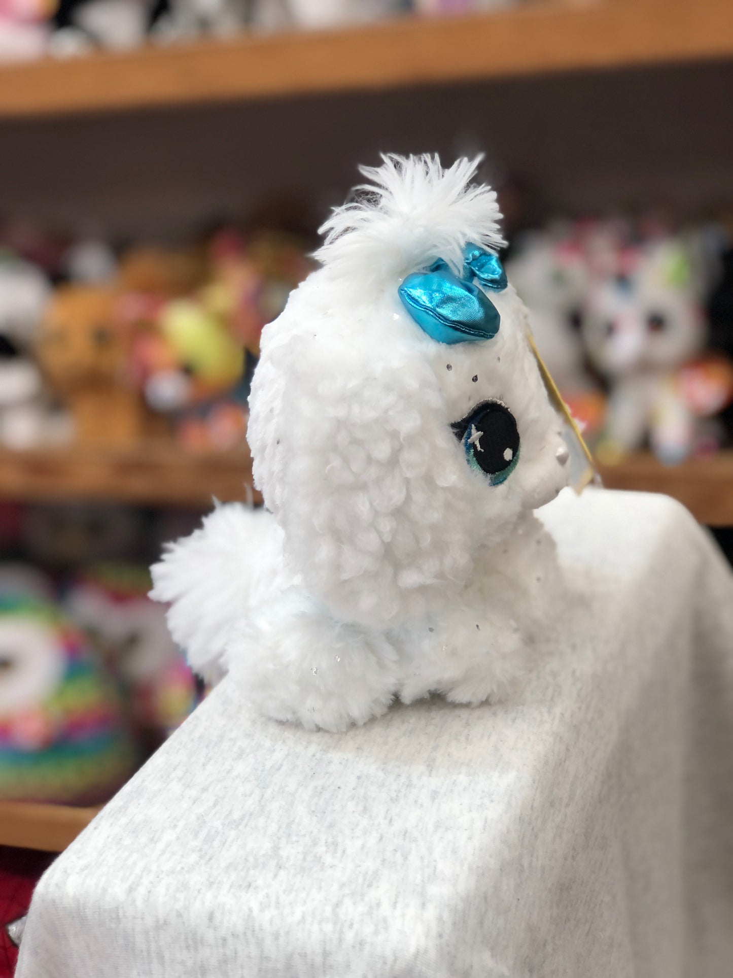 Gund P.Lushes Bianca Blings plush toy: white fluffy dog with sparkly fur, teal eyes, and blue bow, displayed on shelf with other toys