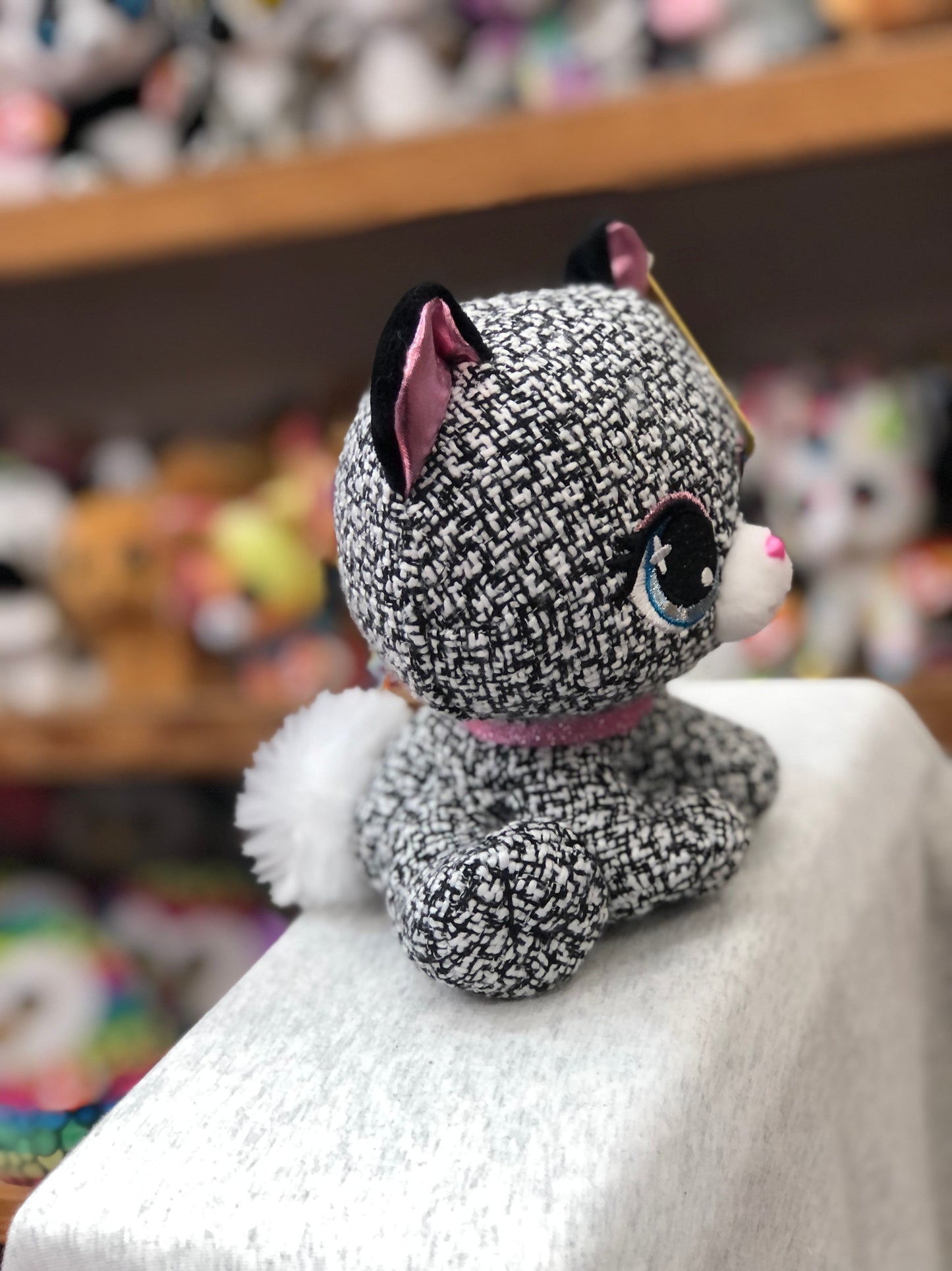 Gund P.Lushes Khloe O'Bearci plush toy with tweed-like fabric, pink ears, blue eyes, and glittery collar, displayed on shelf