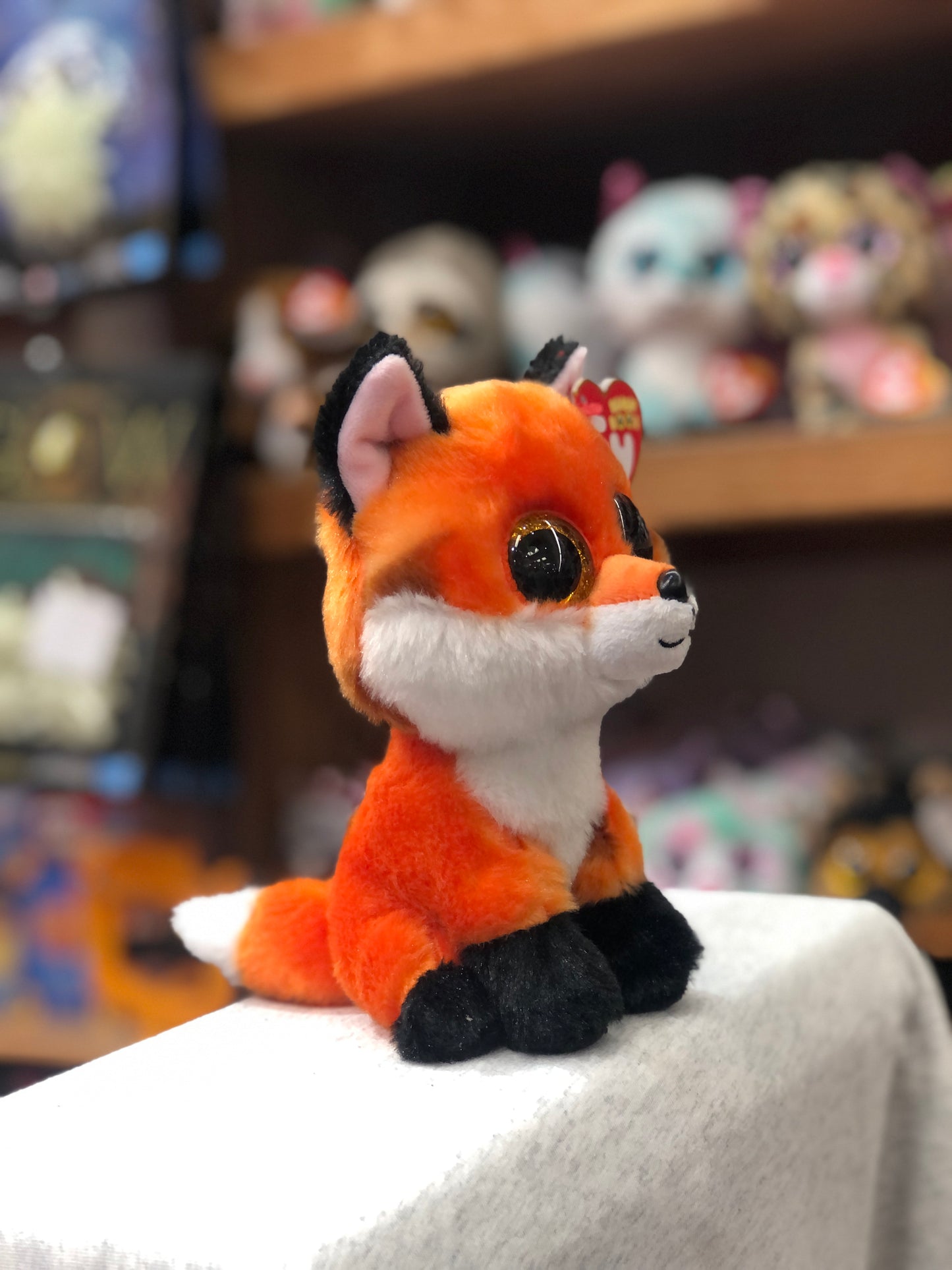 Ty Beanie Boo Meadow orange fox plush toy with large eyes, sitting on display with other stuffed animals in background