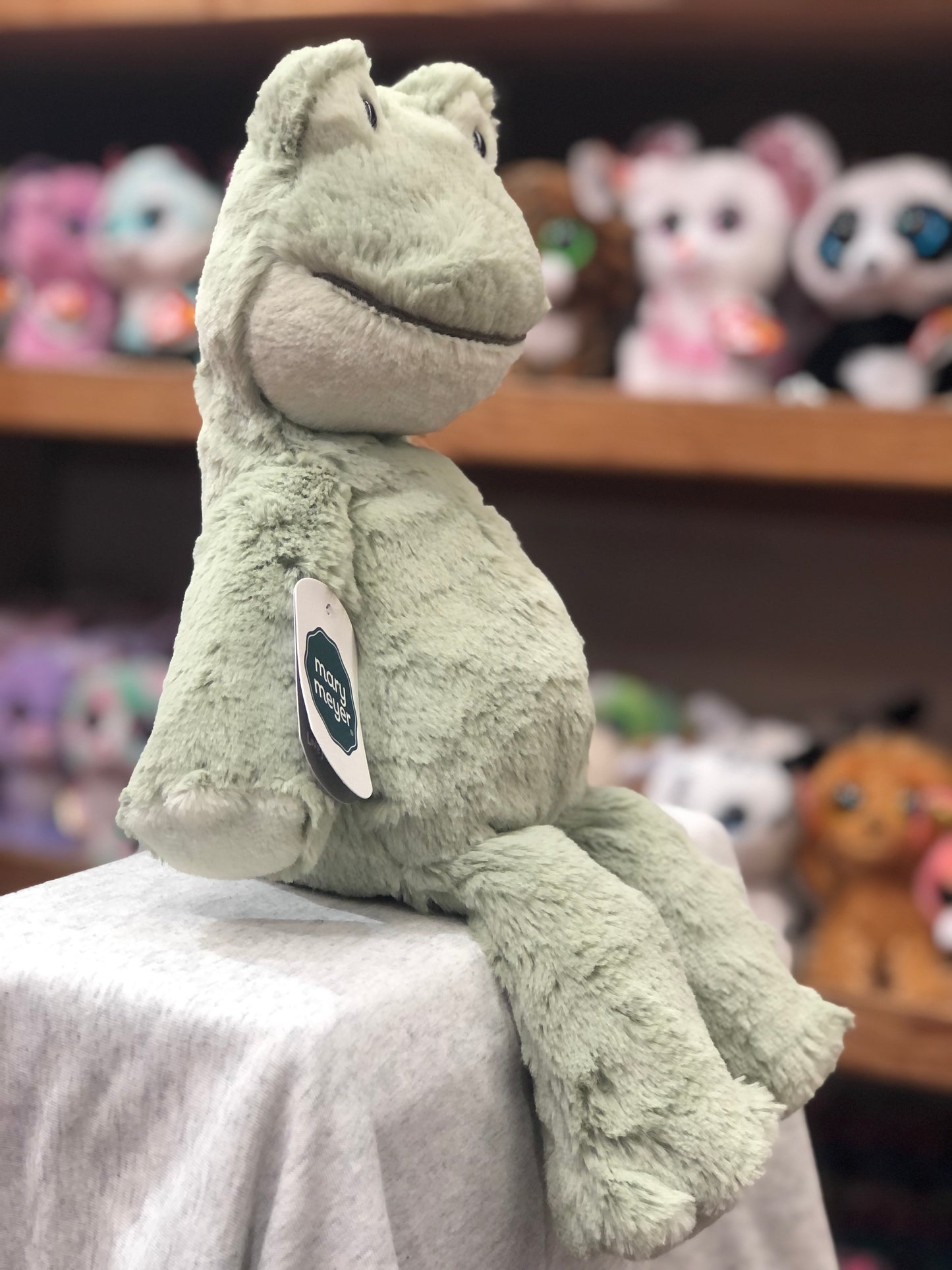 Soft Mary Meyer Marshmallow Mossy Frog plush toy sitting on display, with other stuffed animals visible in background
